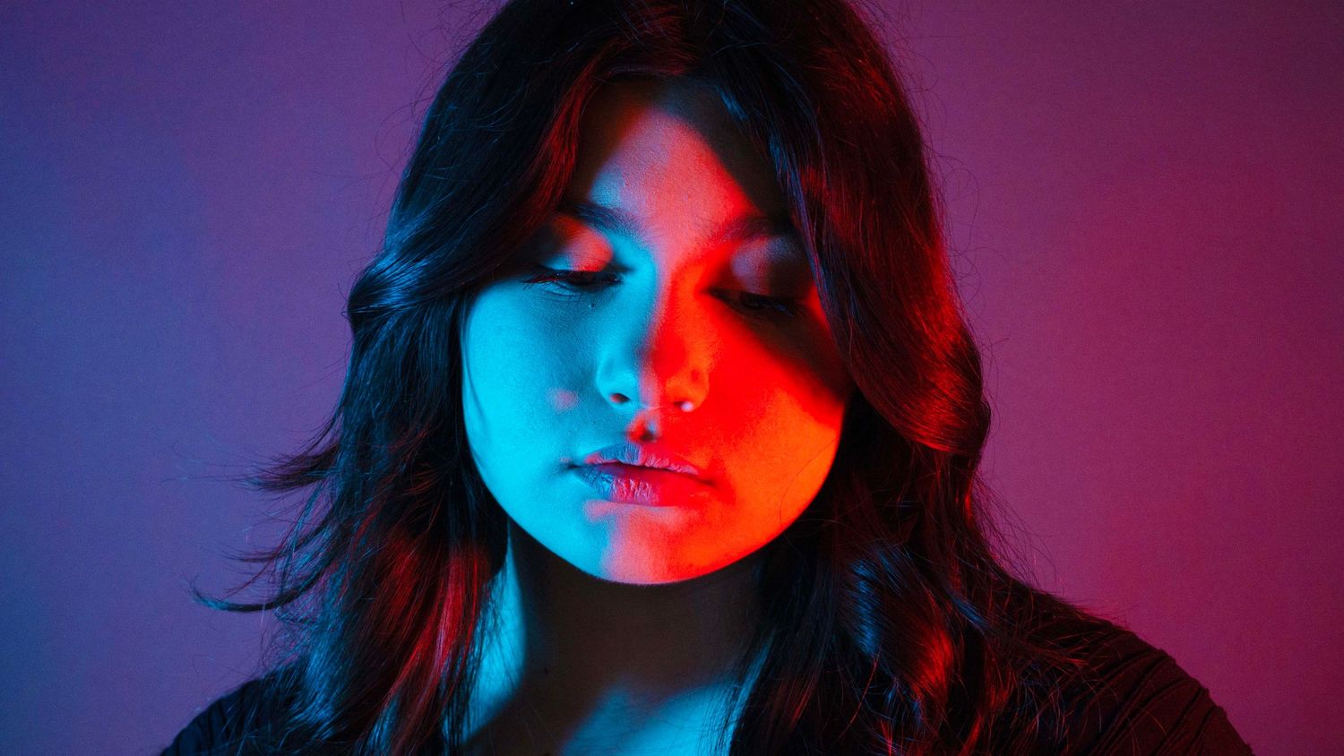 Young woman with dark hair poses under dramatic red and blue neon lighting against a purple background.
