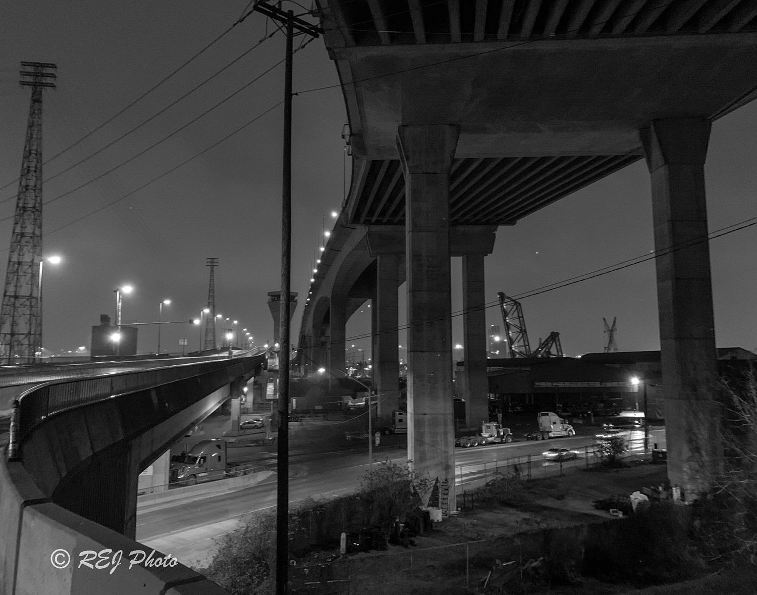 Black and white industrial cityscape at night showing elevated roads and street lights creating moody urban atmosphere.