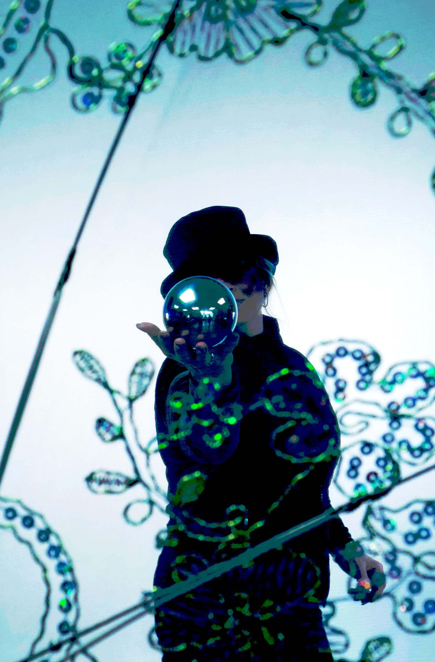 Person in black hat holding reflective orb against glowing teal floral light installation backdrop.