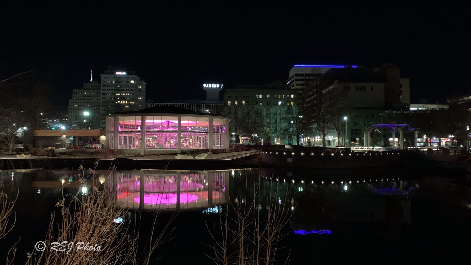 A nighttime cityscape with illuminated buildings and pink lighting reflecting on the waterfront.