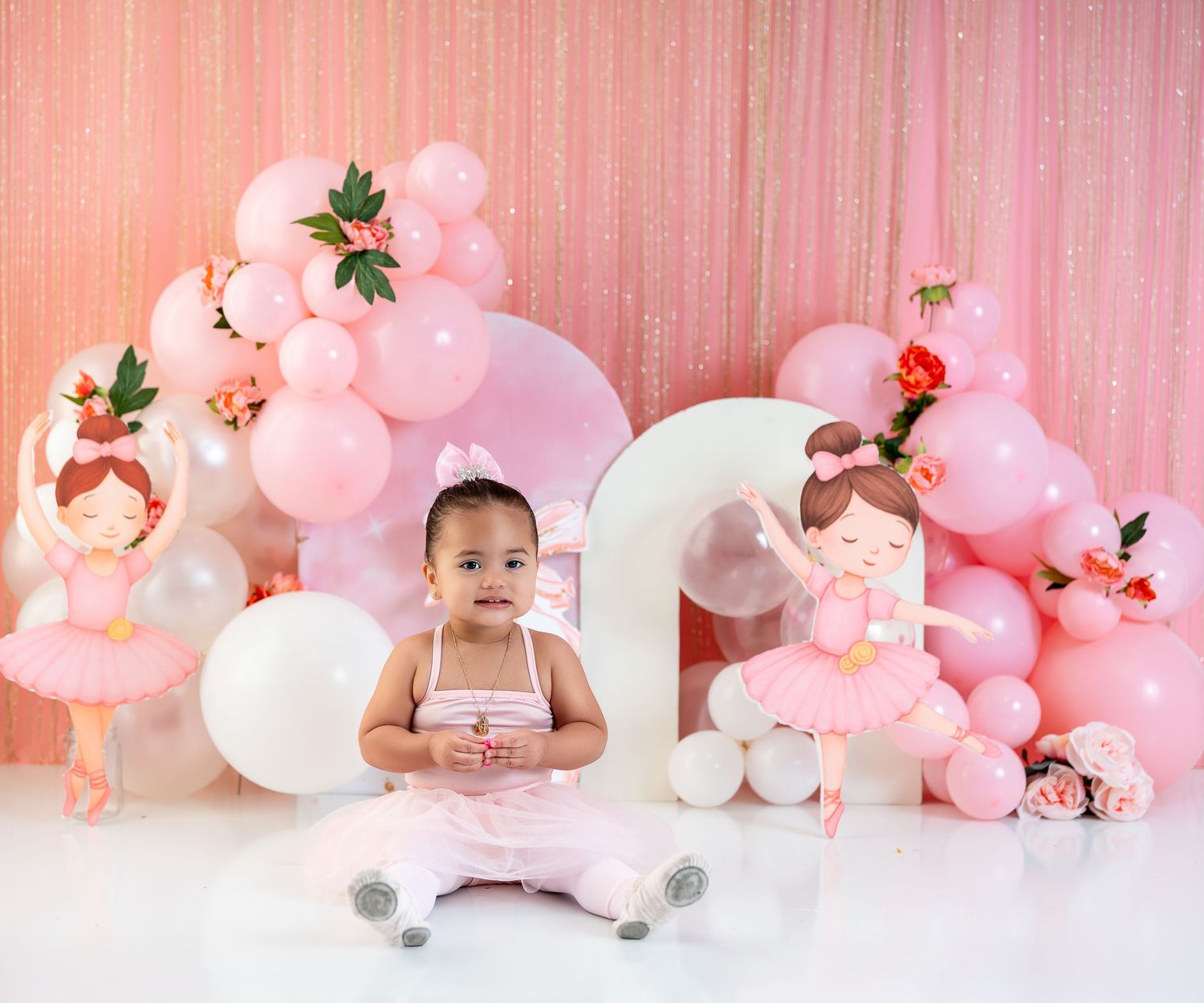 Baby girl in pink tutu sits before a first birthday balloon arch with ballerina cutouts and floral decorations.