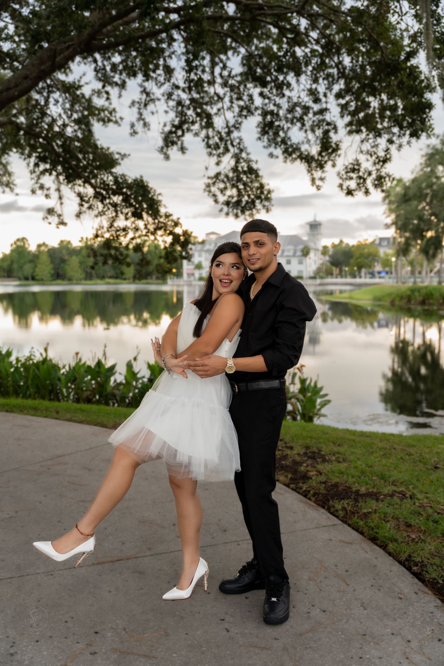 Playful couple in formal wear poses by a lakeside path under tree branches.