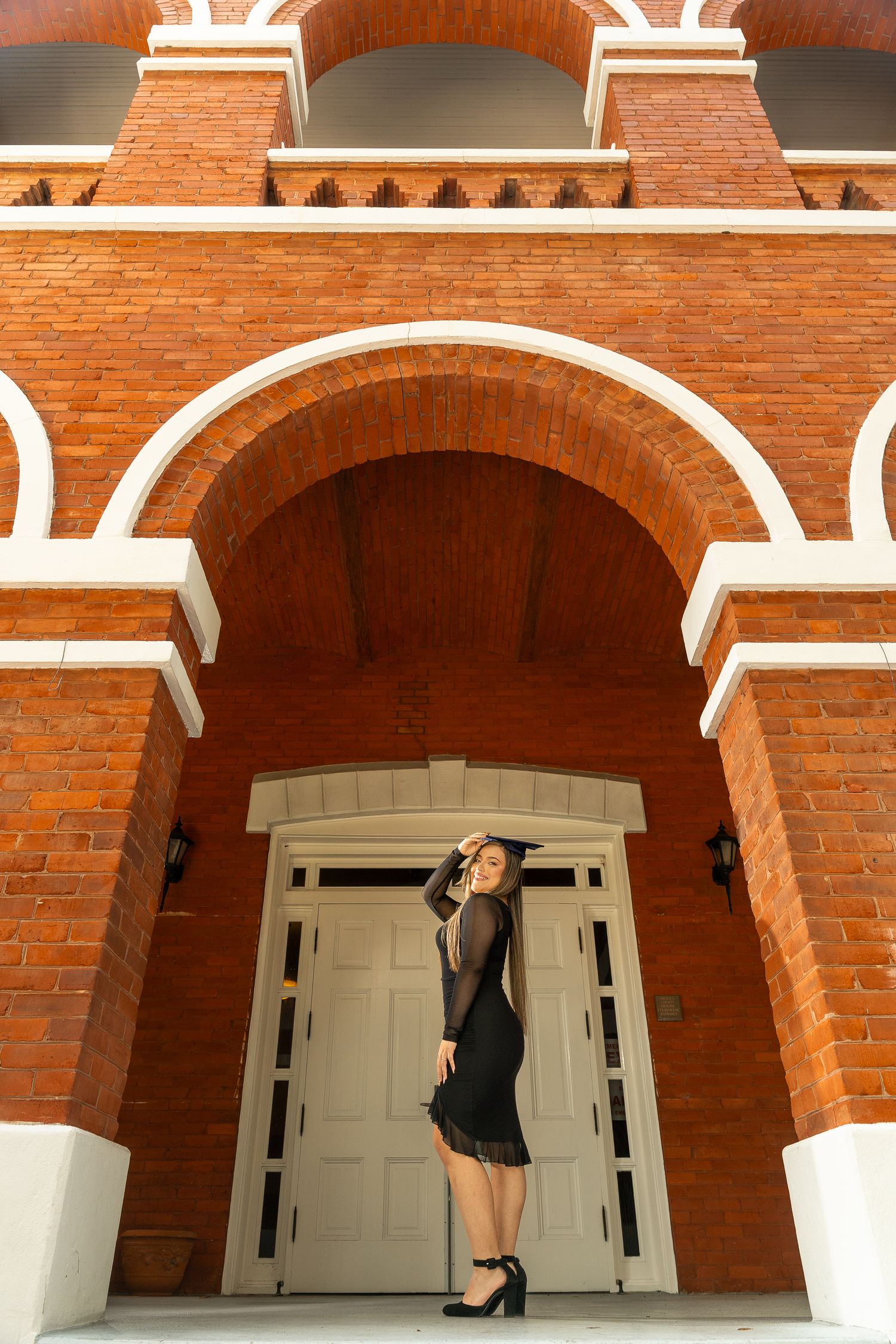 A woman in a black dress poses elegantly in front of an ornate brick archway with white trim and double doors.