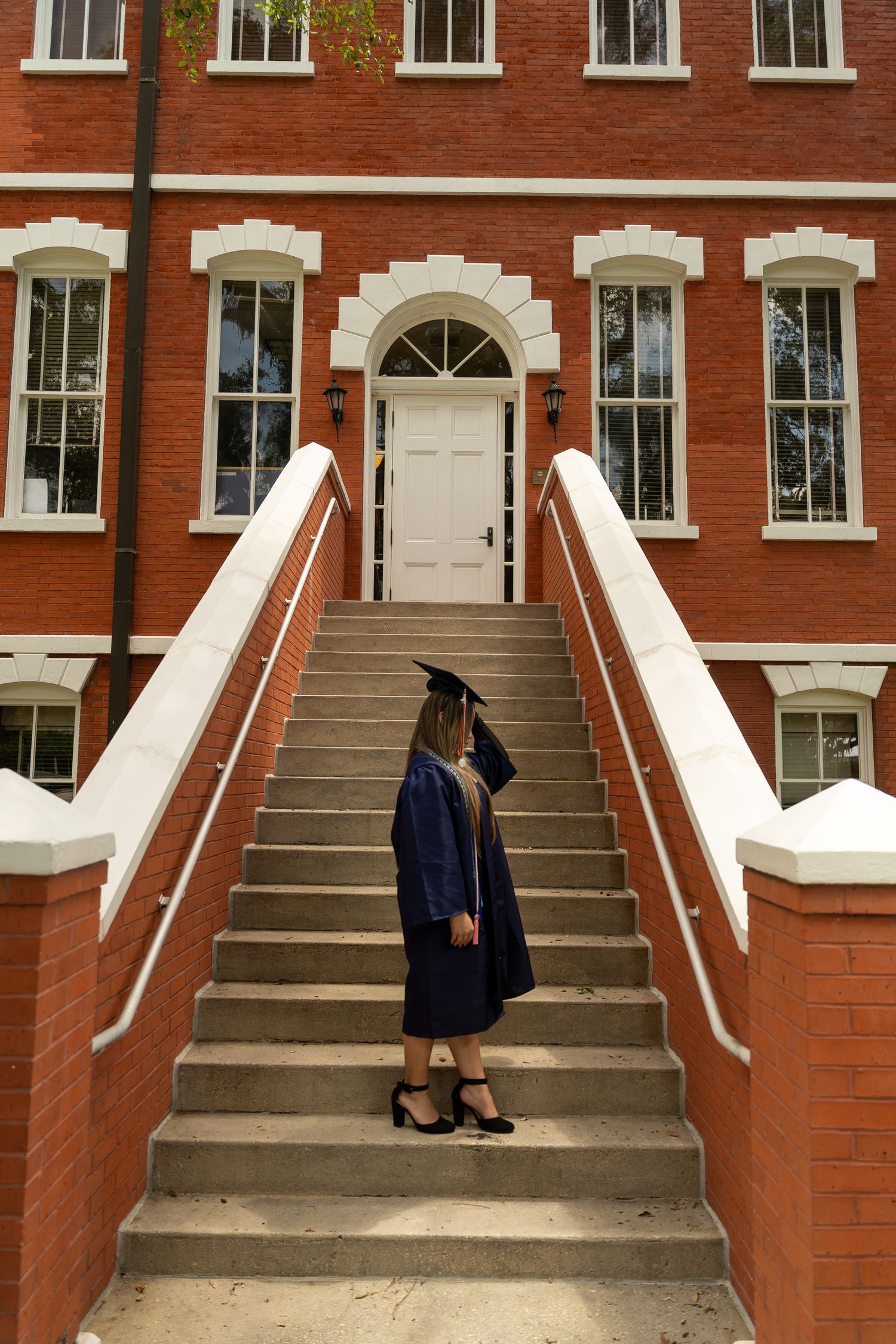 Graduate in cap and gown stands on brick staircase leading to white door of traditional red brick building.