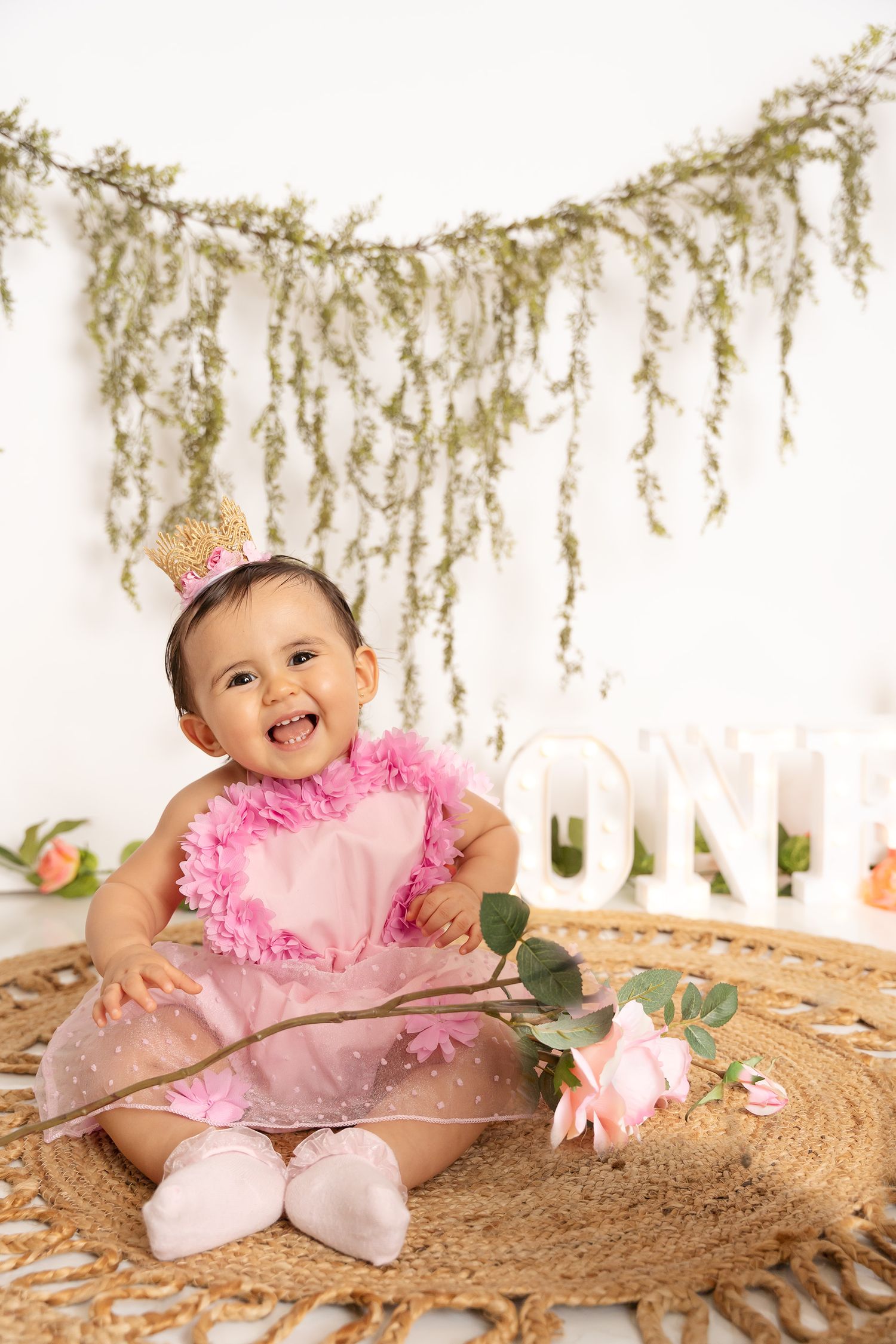 Birthday photoshoot on woven mat with hanging greenery and pink floral accents.