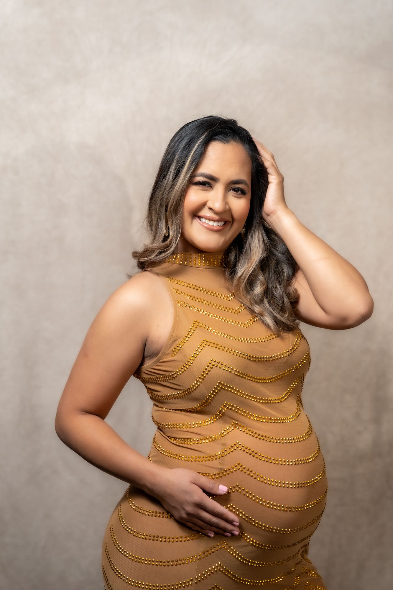 Maternity photoshoot with woman in golden dress against neutral background.