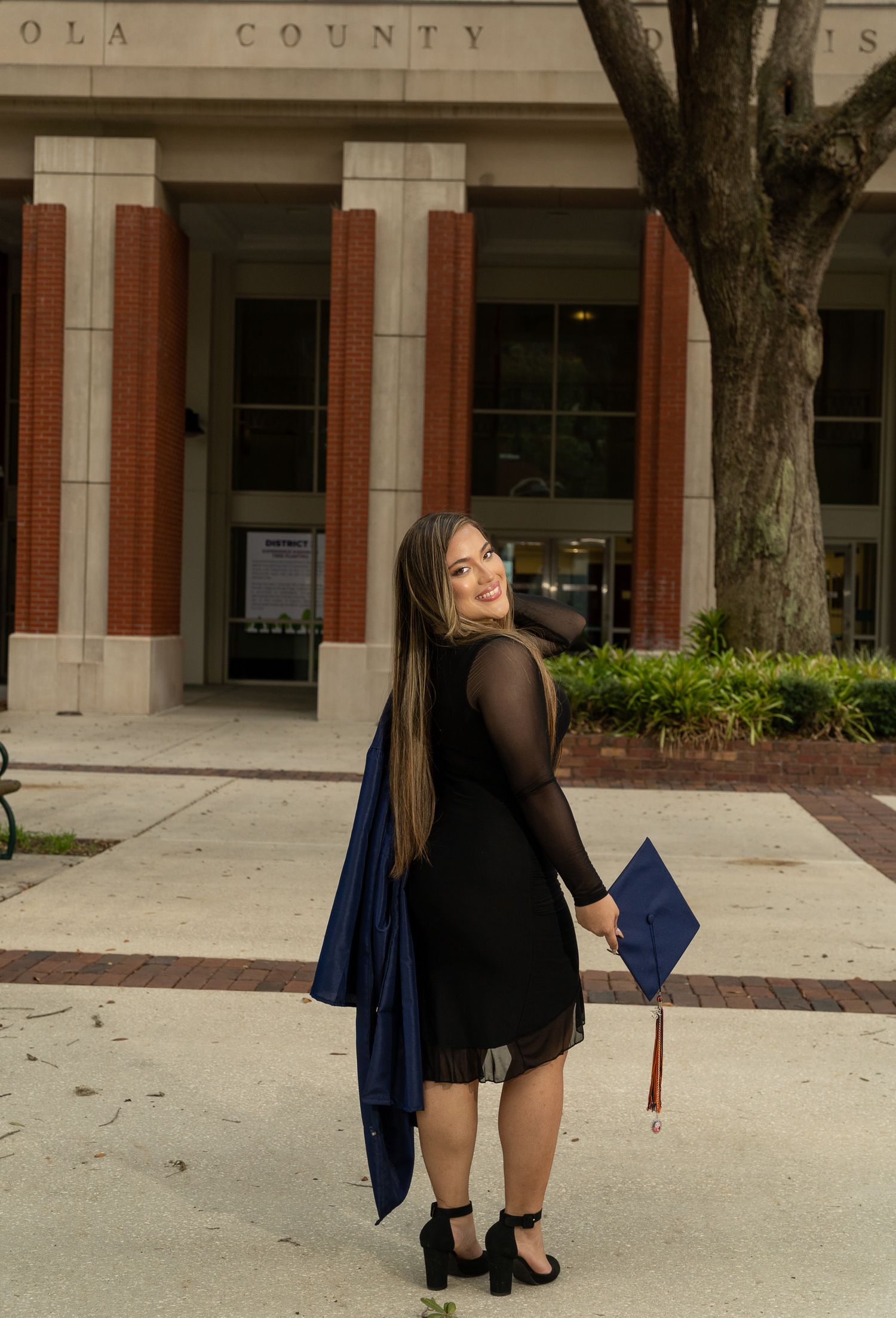 A graduate in a black dress and navy blue cap and gown stands outside a county building during commencement.