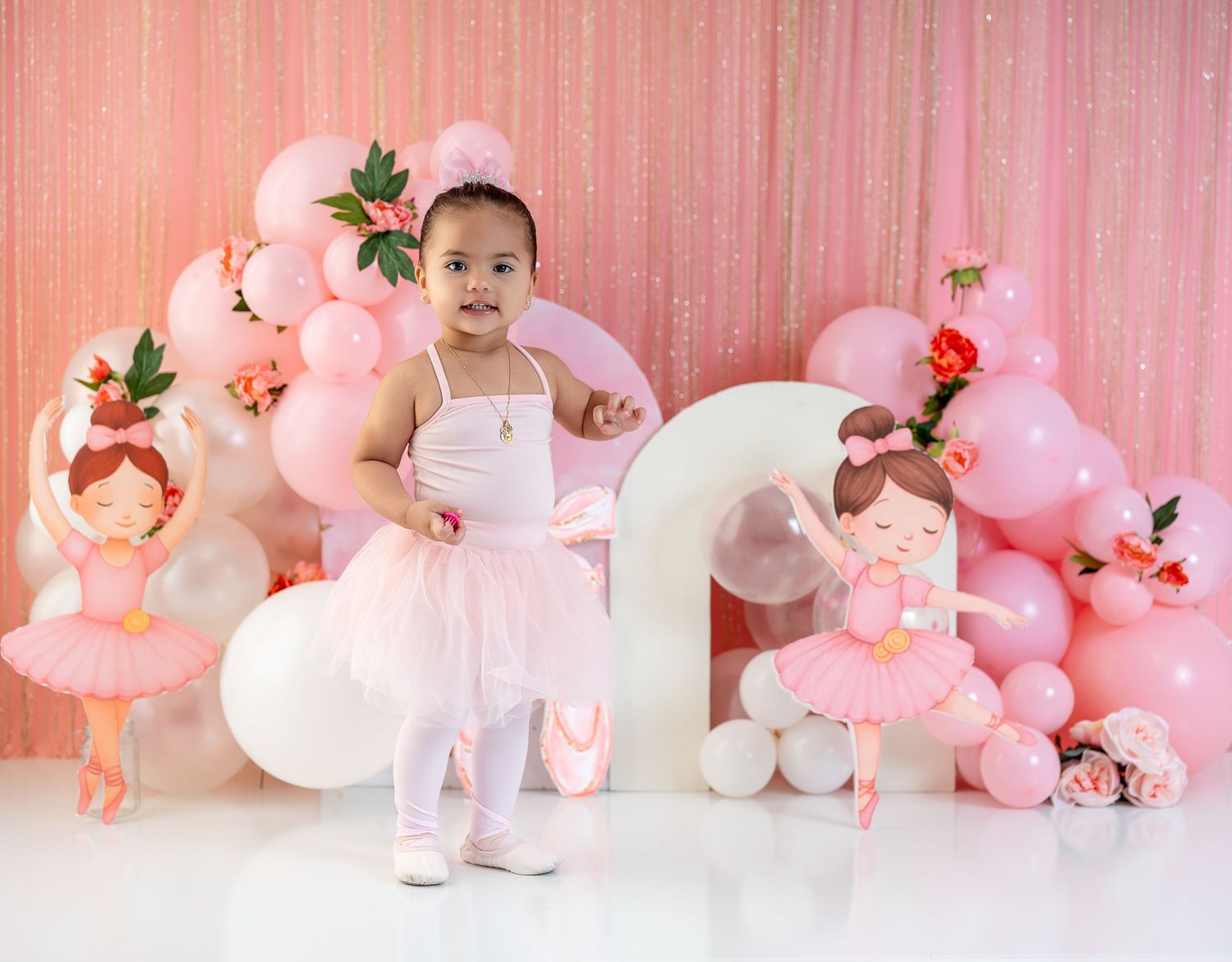 Adorable toddler in pink ballerina tutu poses with balloon arch and ballerina cutout decorations for her 2nd birthday.