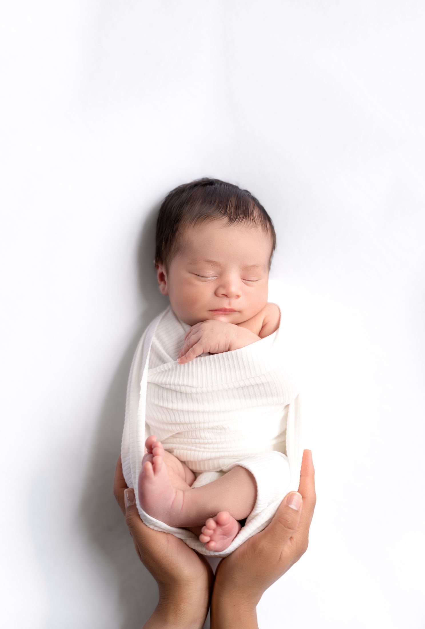 Minimalist newborn photography with baby wrapped in white against clean background.