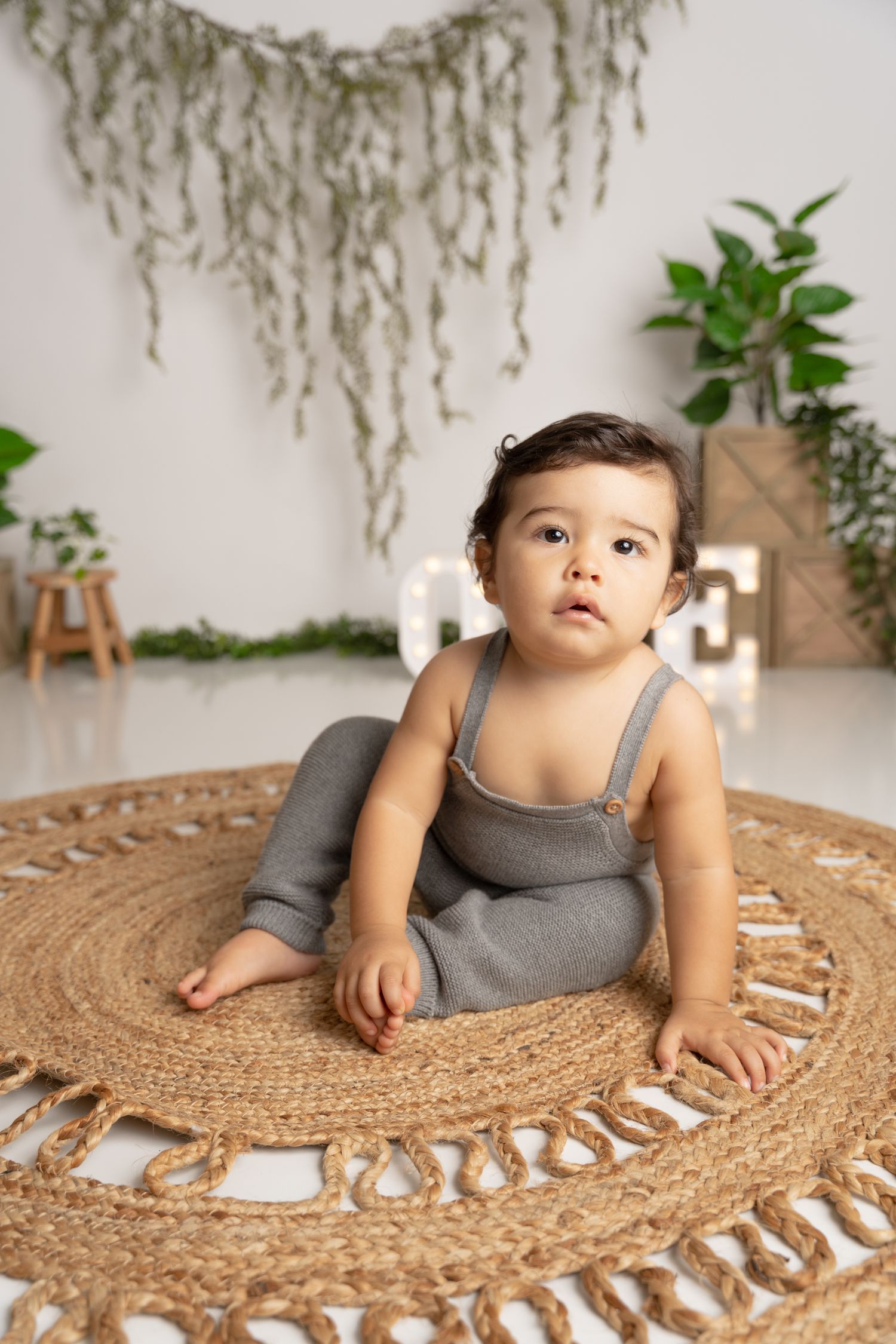 Baby in grey romper crawls on round woven jute rug with decorative tassels against white wall with hanging greenery.