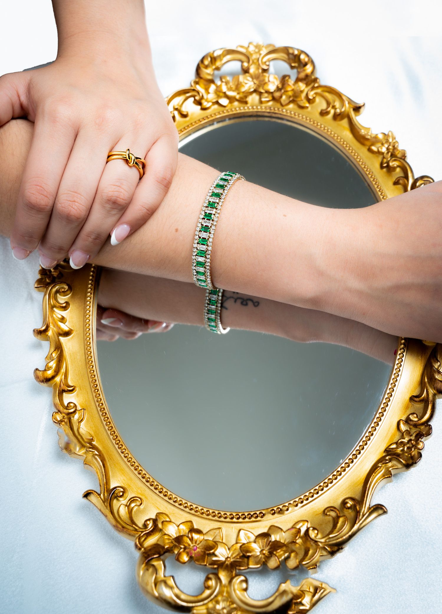 Hand displaying jewelry against ornate gold mirror with emerald bracelet and rings.