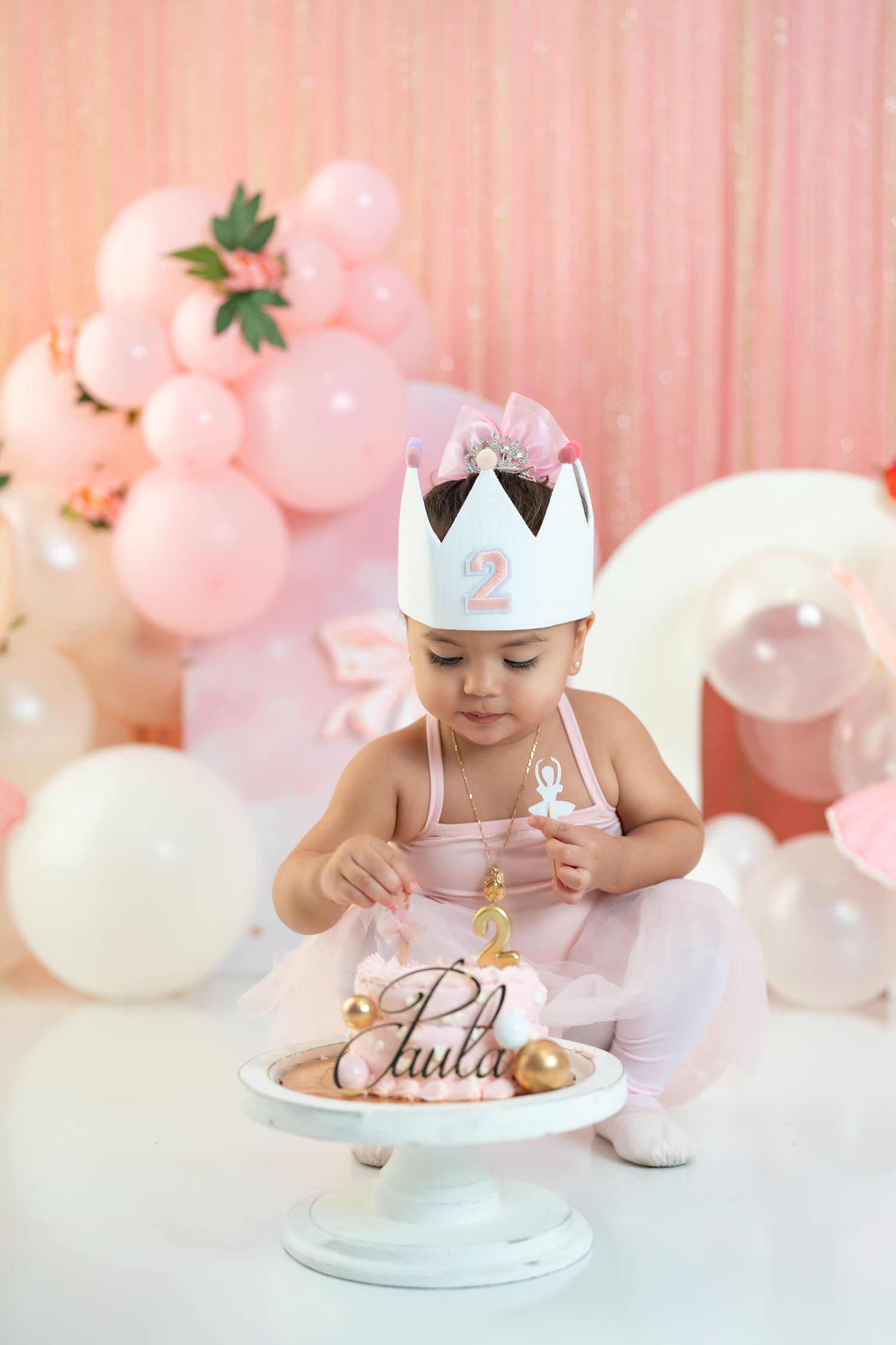 Toddler girl in pink tutu and crown hat reaches for her second birthday cake named Paula with gold decorations.