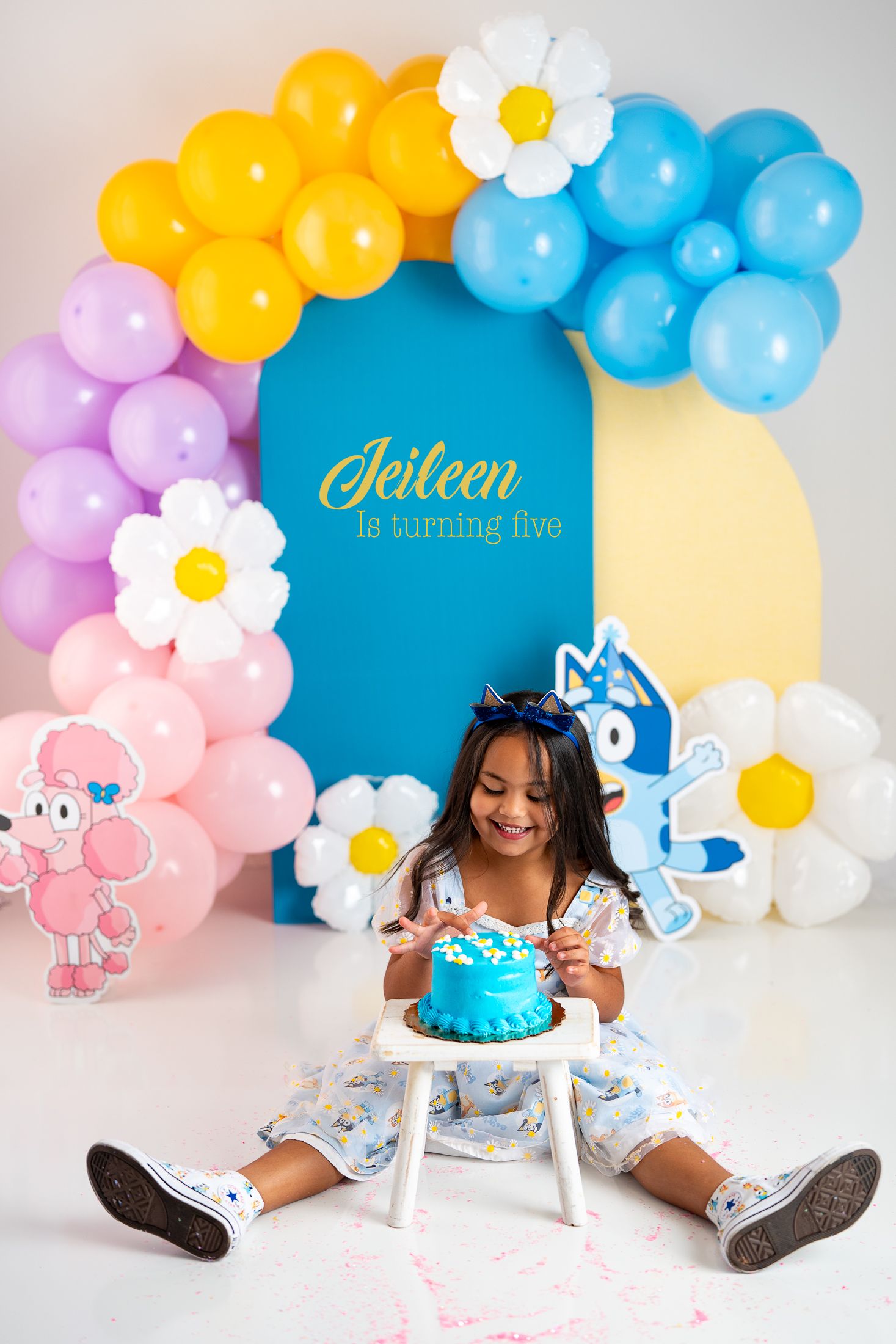 Birthday setup with colorful balloon arch and blue cake against pastel backdrop.