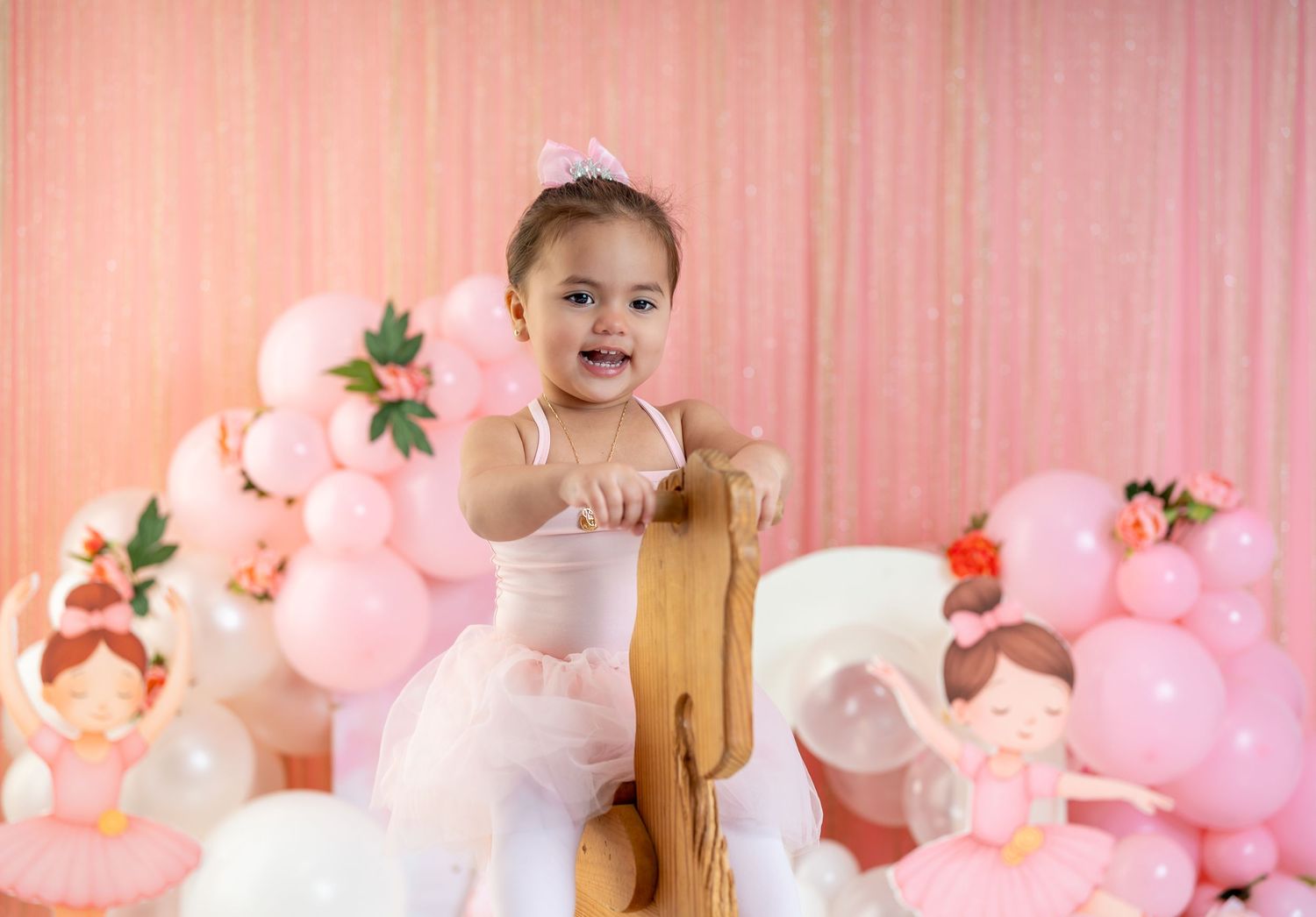 Toddler girl in pink tutu and tiara smiles at first birthday party with balloon decorations.