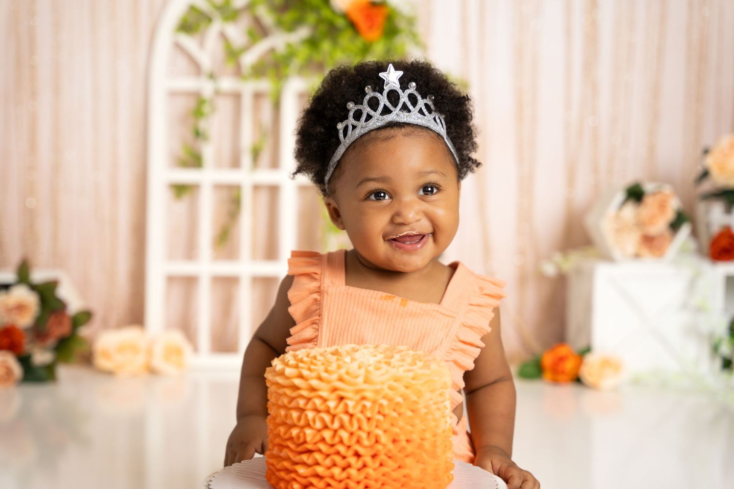 Festive birthday setup with orange ruffle cake and floral decorations against white backdrop.