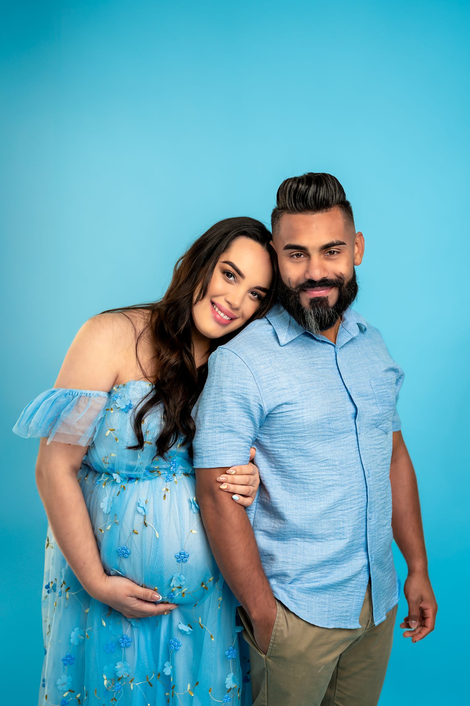 Couple in coordinating light blue outfits posing against turquoise backdrop for maternity photos.