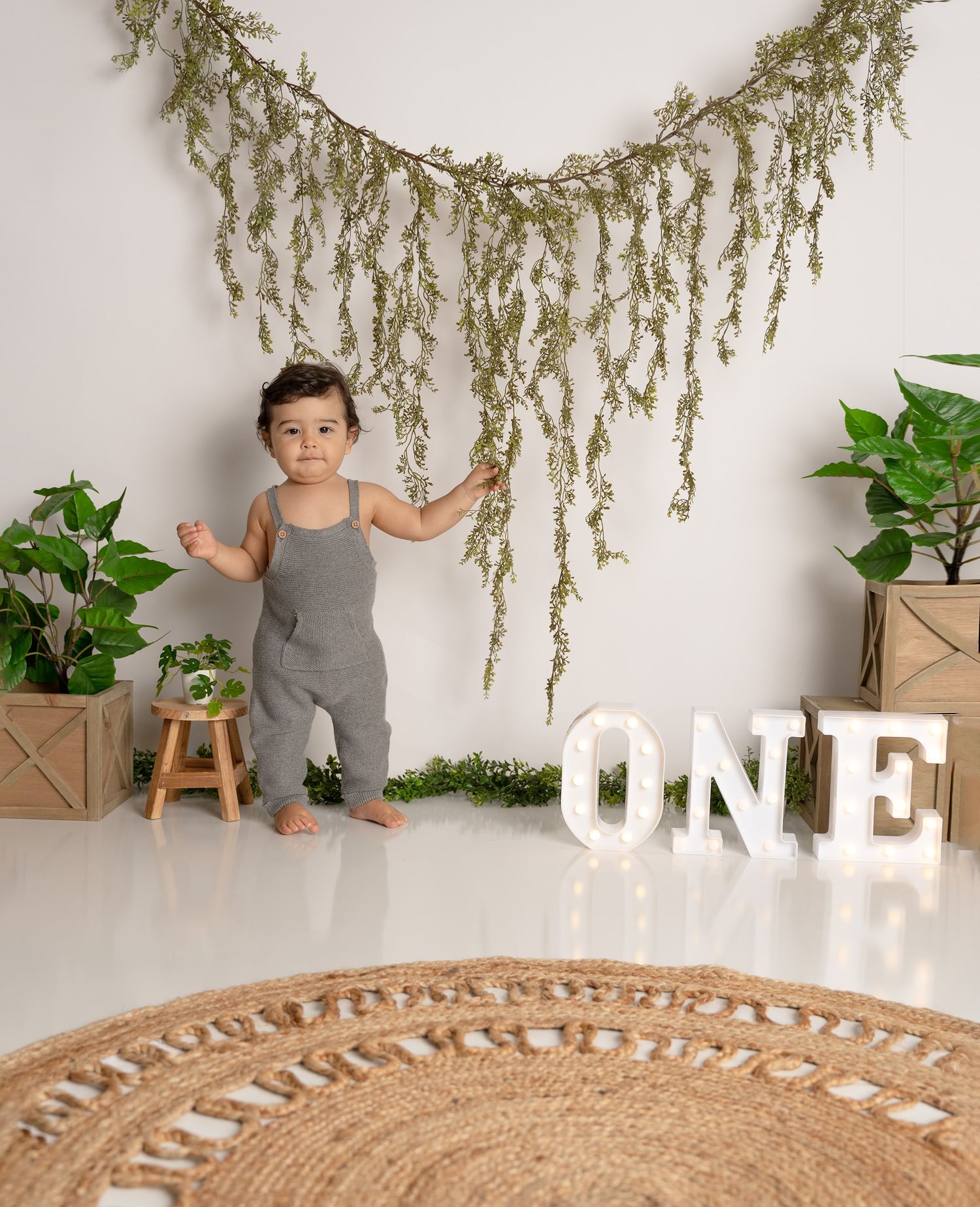 Minimalist birthday setup with hanging greenery and illuminated ONE sign on round jute rug.
