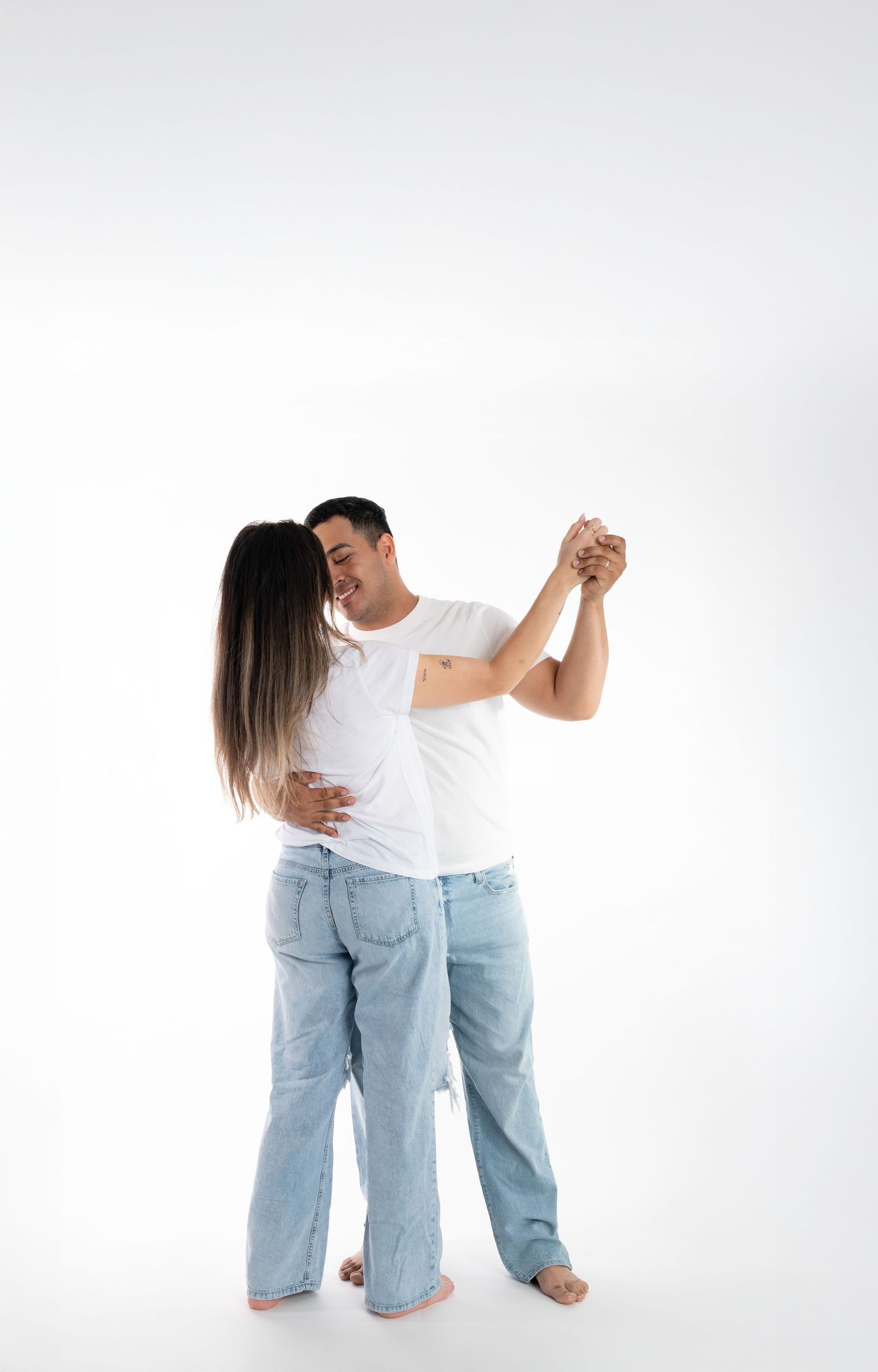 A couple in matching white t-shirts and light denim jeans dance together barefoot against a white background.