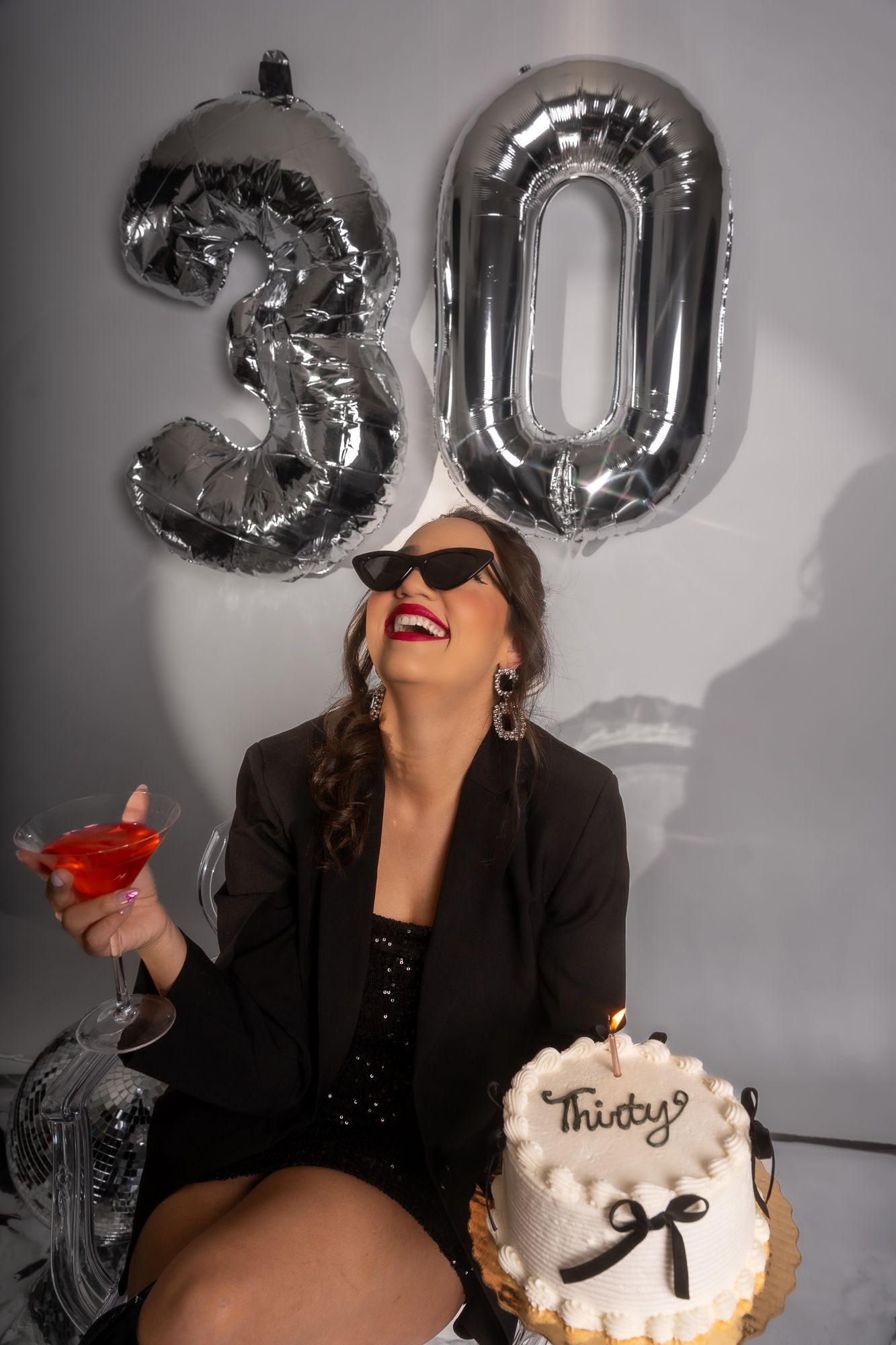 Celebratory scene with silver number balloons, red cocktail, and white birthday cake against grey backdrop.