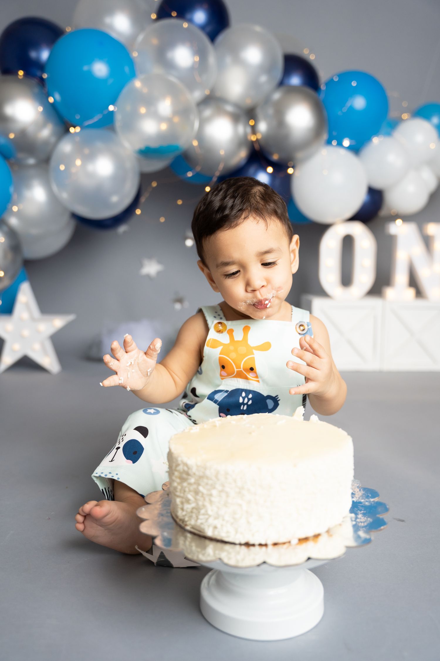 White cake smash photography session with blue and silver balloon arch backdrop and sparkling lights.