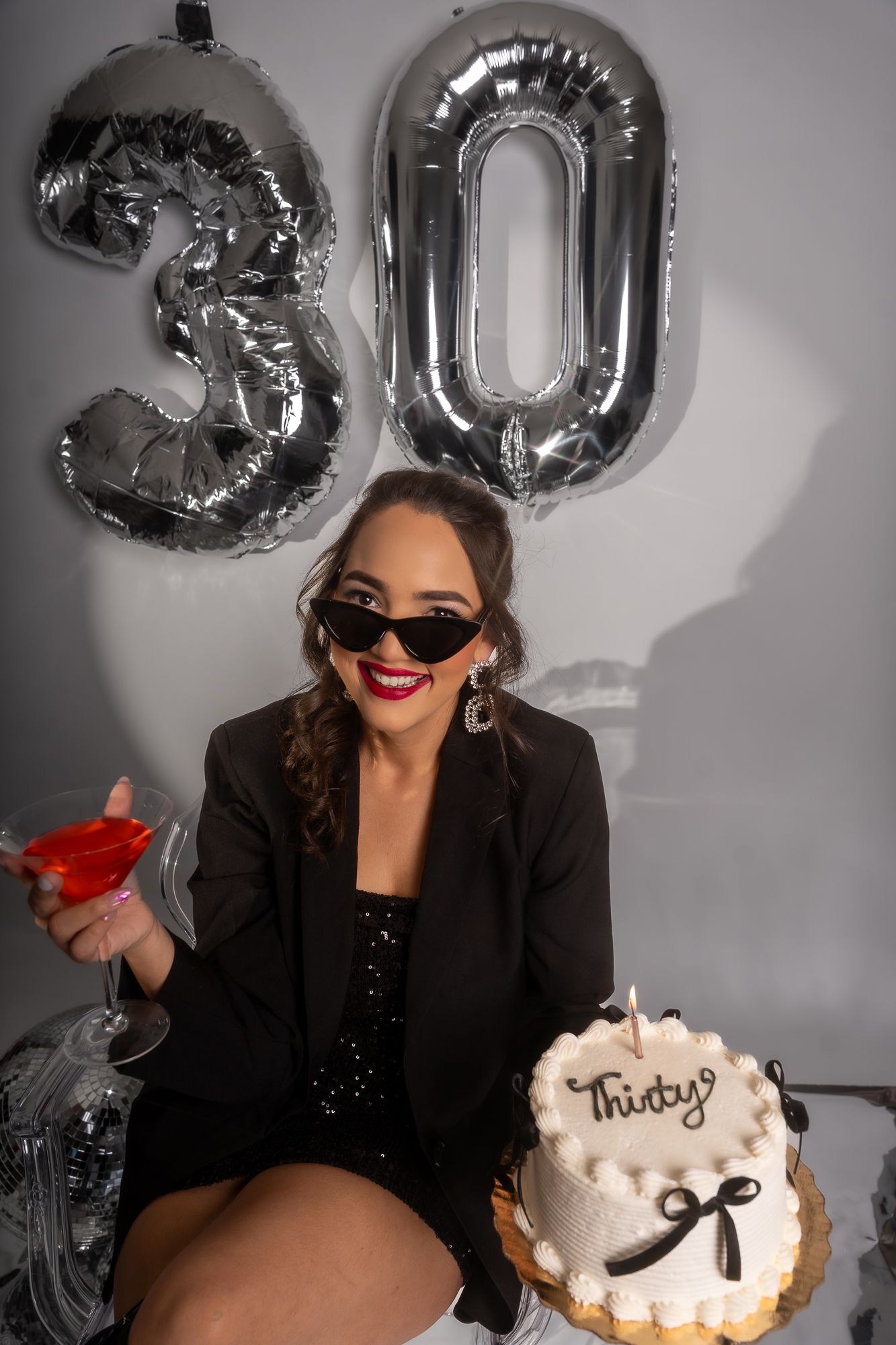 Glamorous birthday scene with silver '30' balloons, red martini and white cake against gray backdrop.