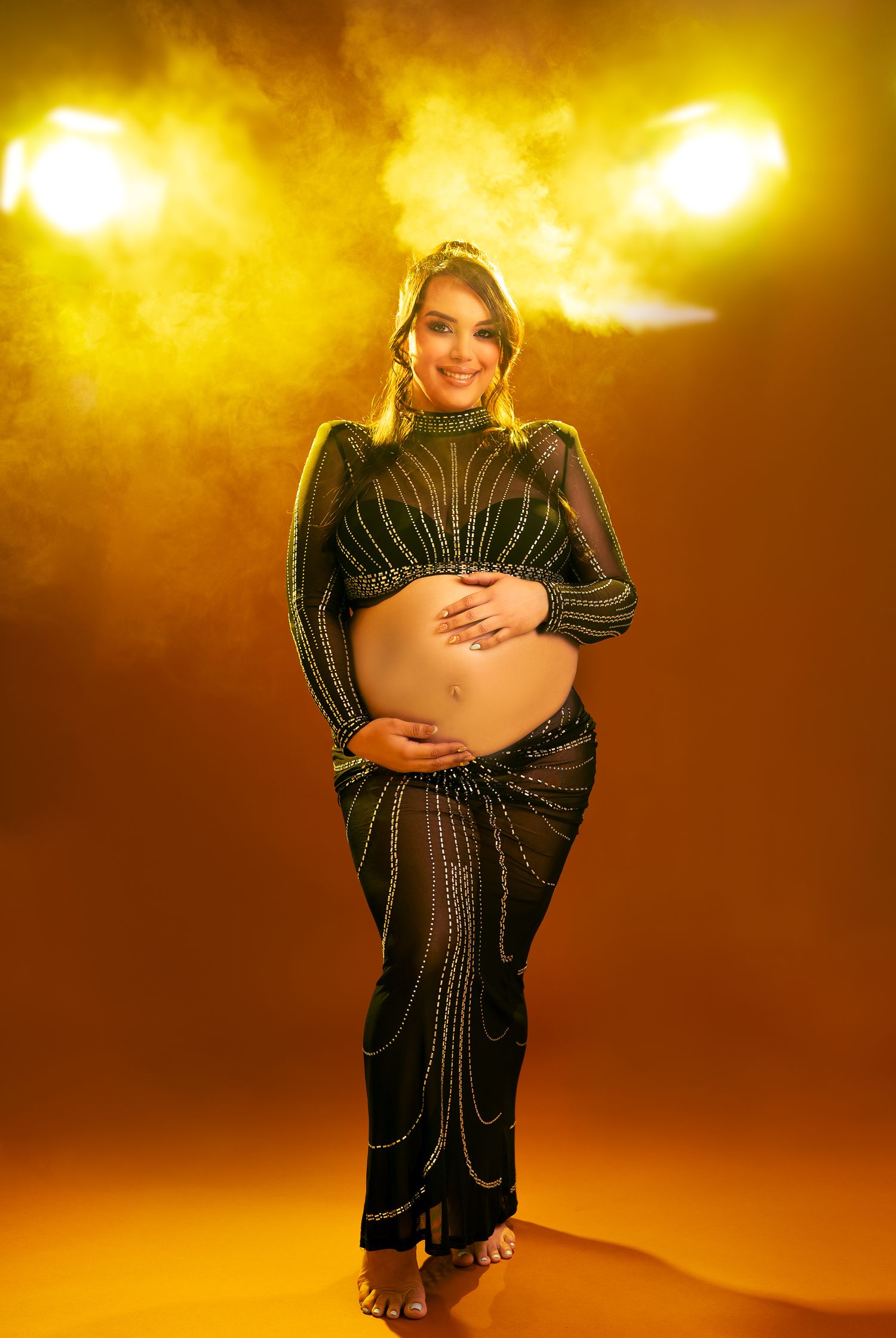 A pregnant woman in a black sequined dress poses against a warm golden background in a maternity photoshoot series.