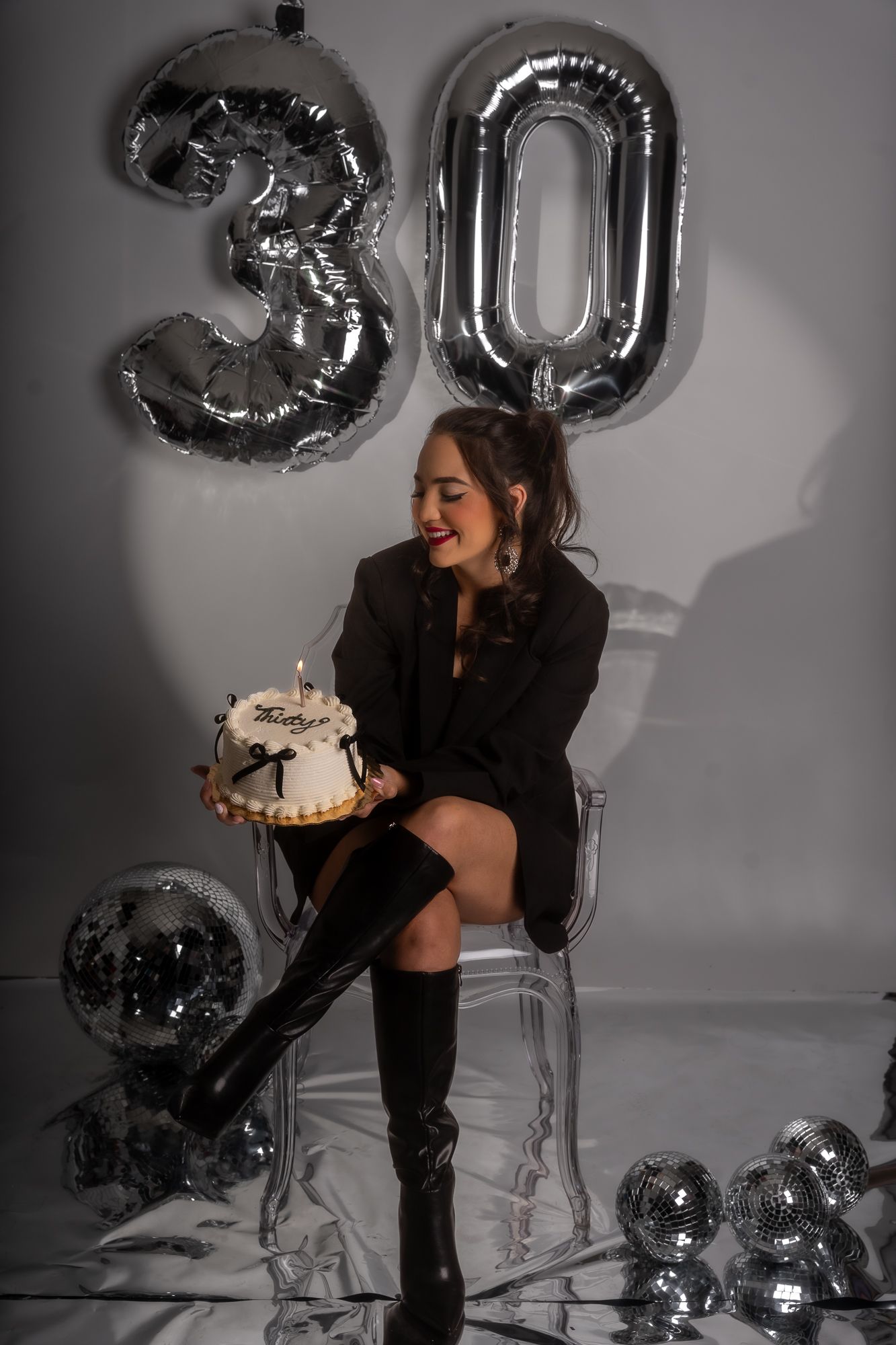 A 30th birthday celebration setup with silver balloons and a white cake on a clear chair against a gray backdrop.