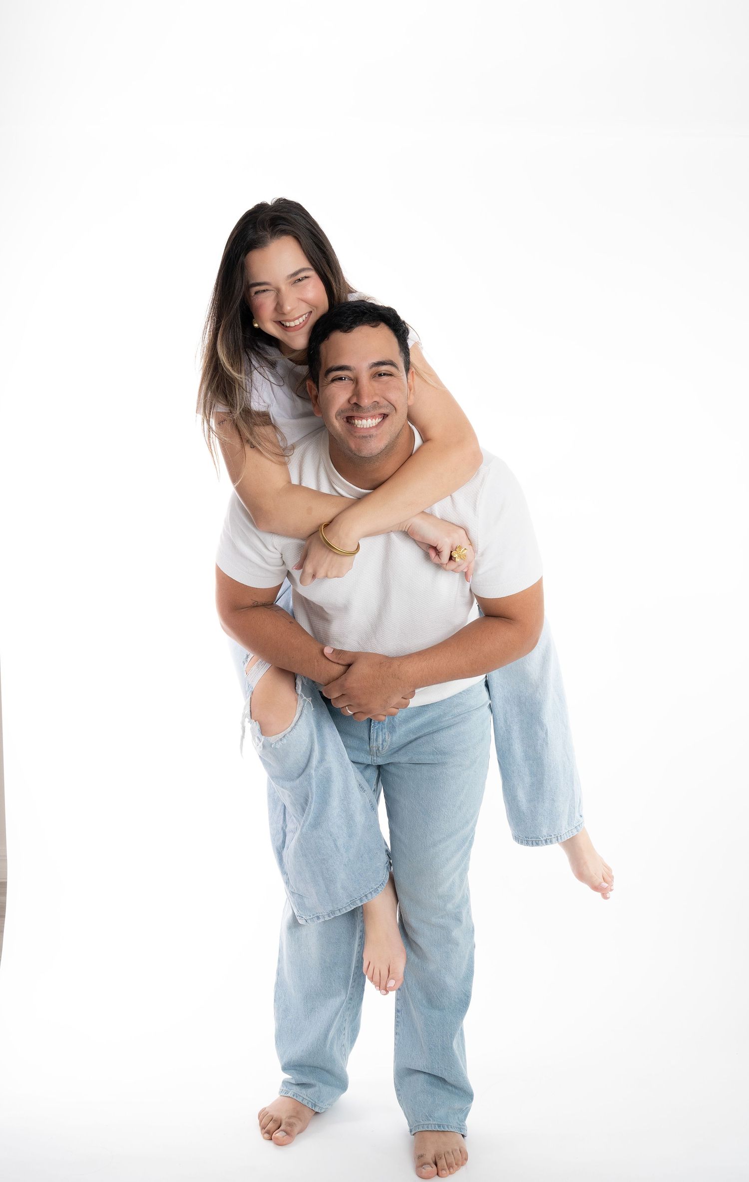 A playful piggyback pose of a couple in matching white shirts and denim against a white backdrop.