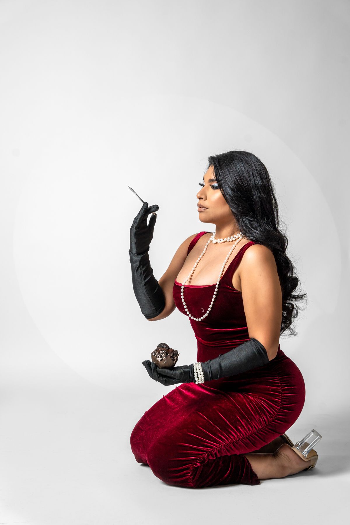Woman in red velvet dress and long gloves posing elegantly with pearl necklace.