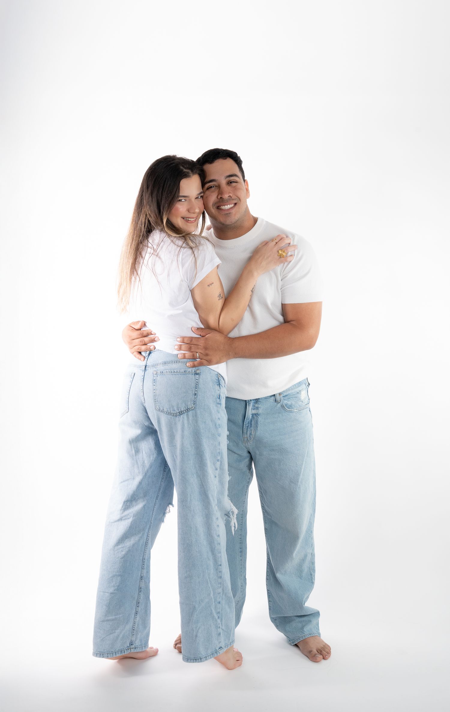 A romantic couple in casual white shirts and jeans embrace while standing against a white backdrop.