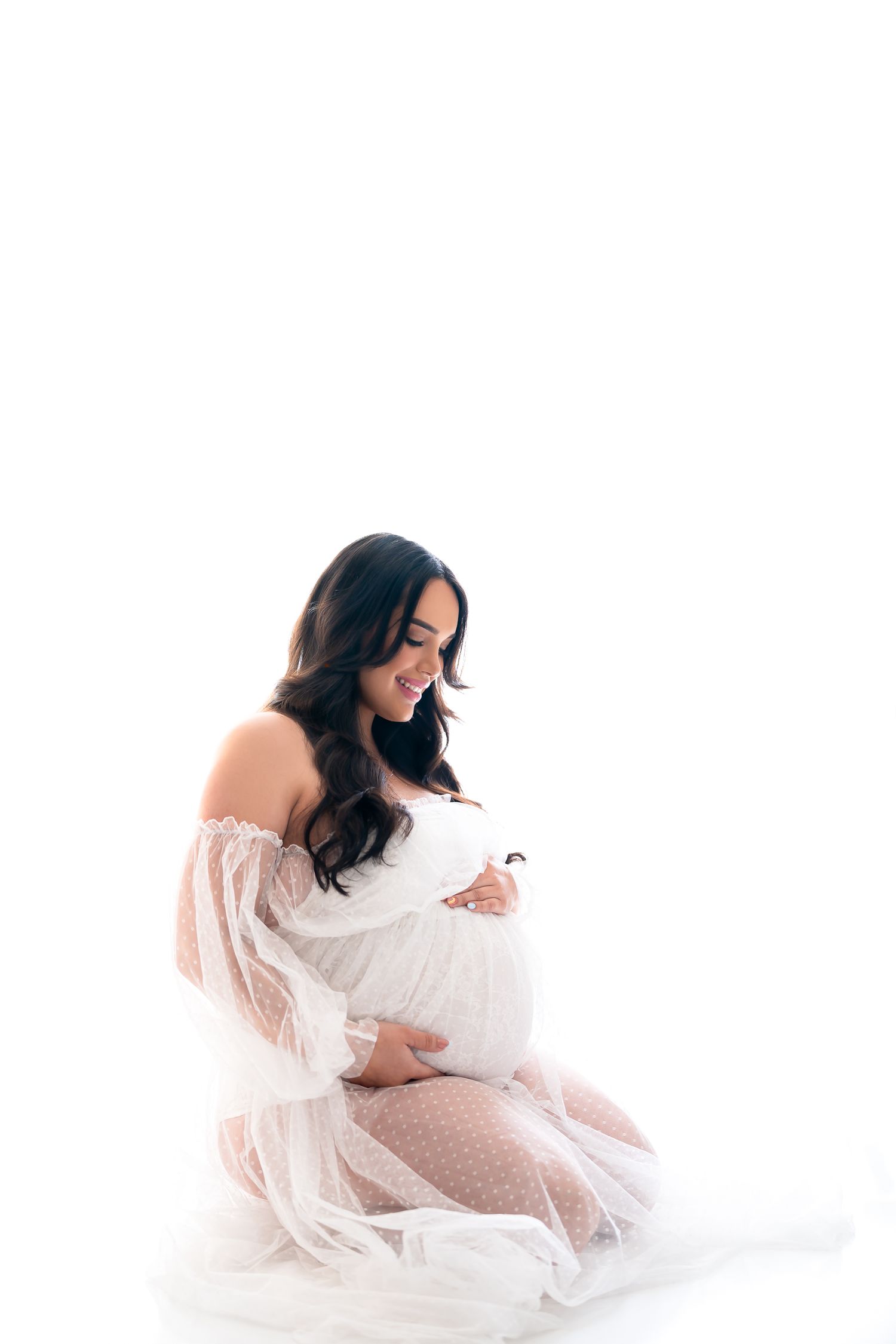 A person in a flowing white lace dress poses gracefully while kneeling on a white background.