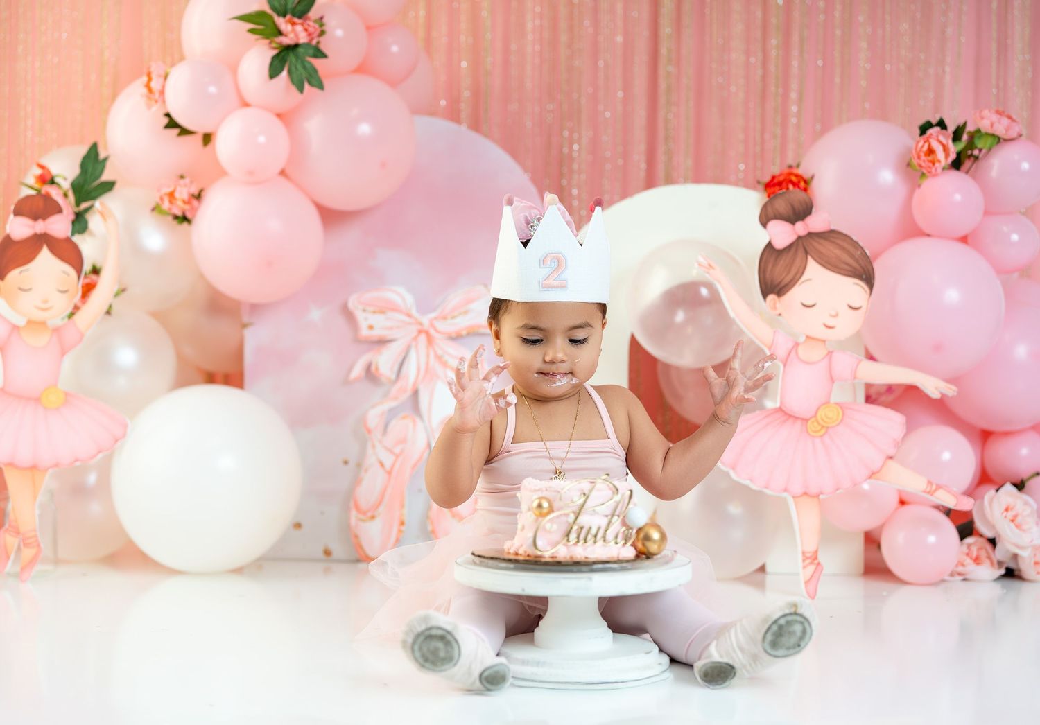 Baby girl in pink tutu smashes first birthday cake surrounded by pink balloon arch and ballerina decorations.