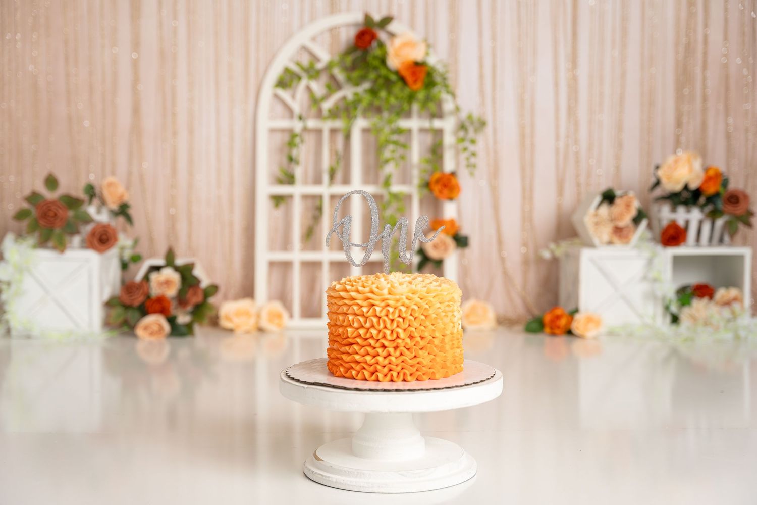 Orange ruffle cake displayed on white cake stand with floral arrangements in background.