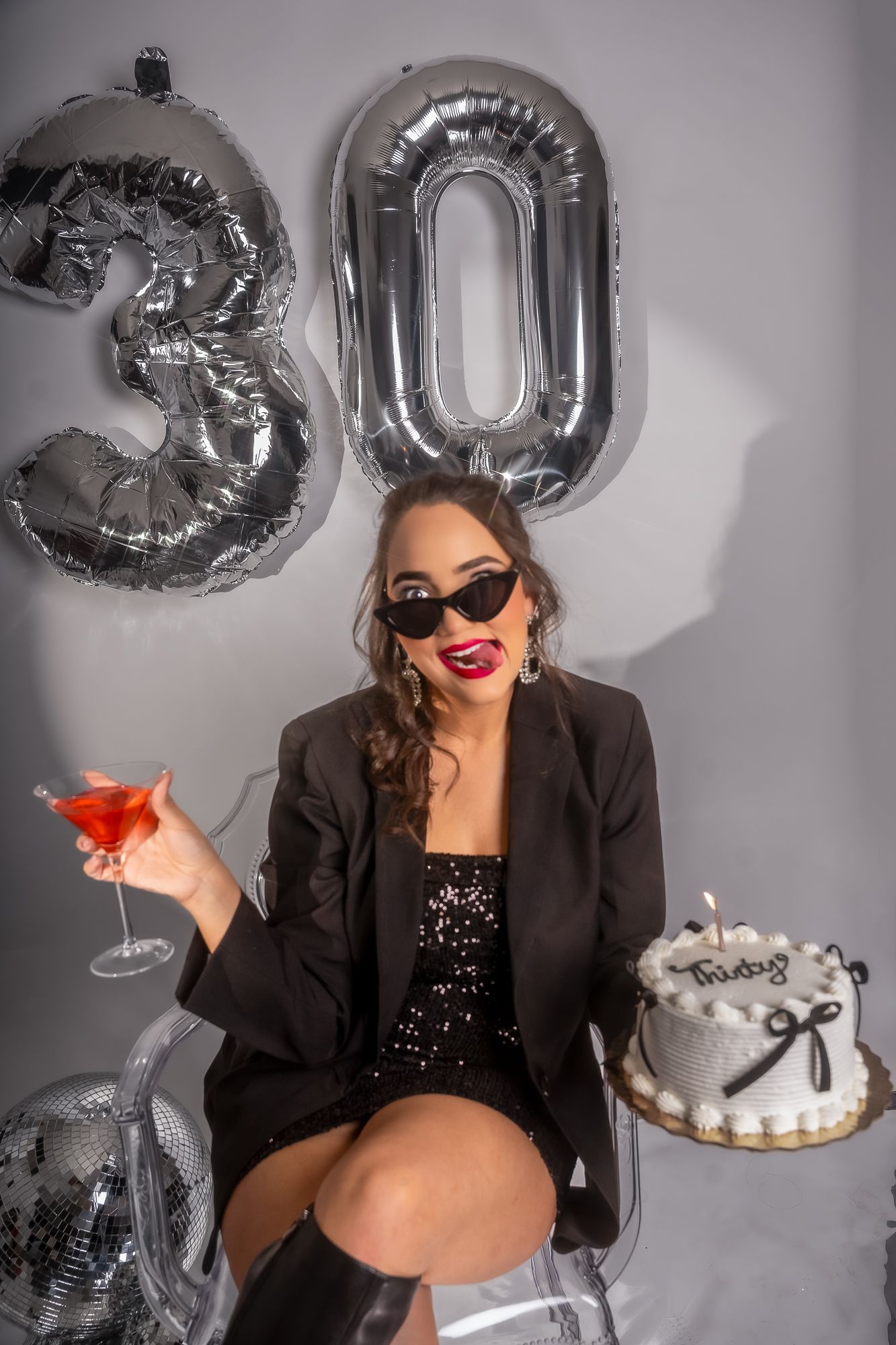 Glamorous birthday setup with silver number balloons, red cocktail, and white cake on clear chair.