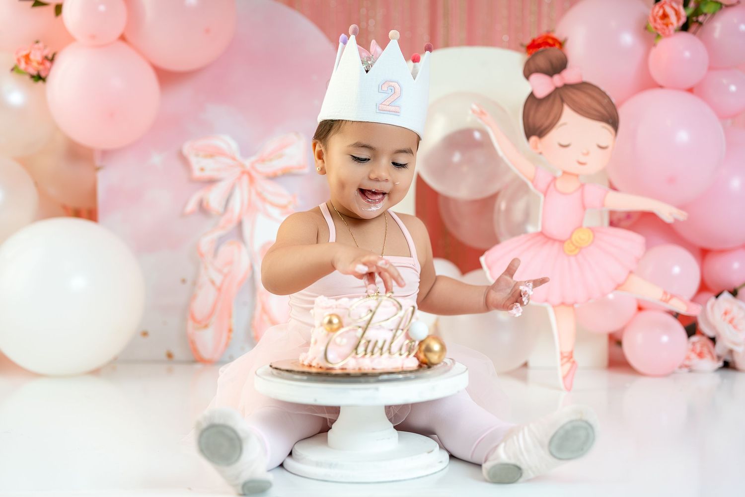 Baby girl in a crown smiles at her first birthday cake surrounded by pink balloons and ballerina decorations.
