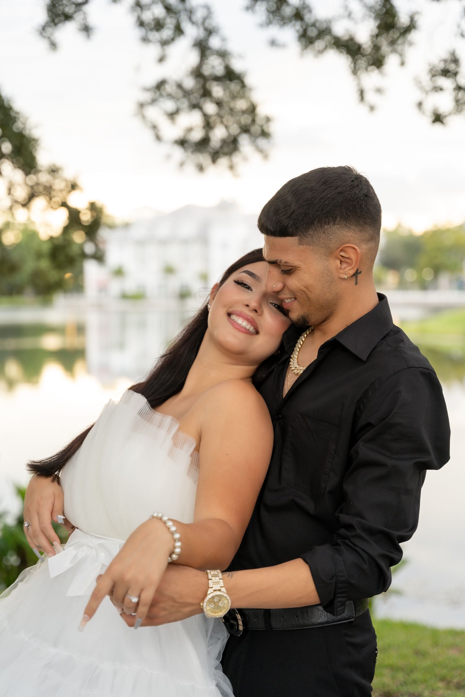 Romantic couple embraces outdoors in formal attire against a scenic lake backdrop at sunset.