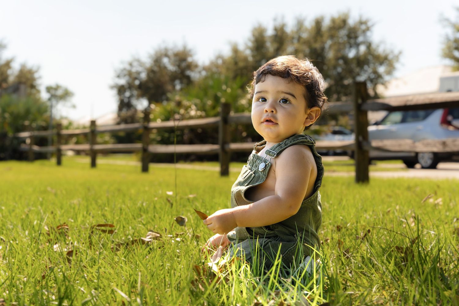 Young child lying in green grass field on sunny day with trees in background.