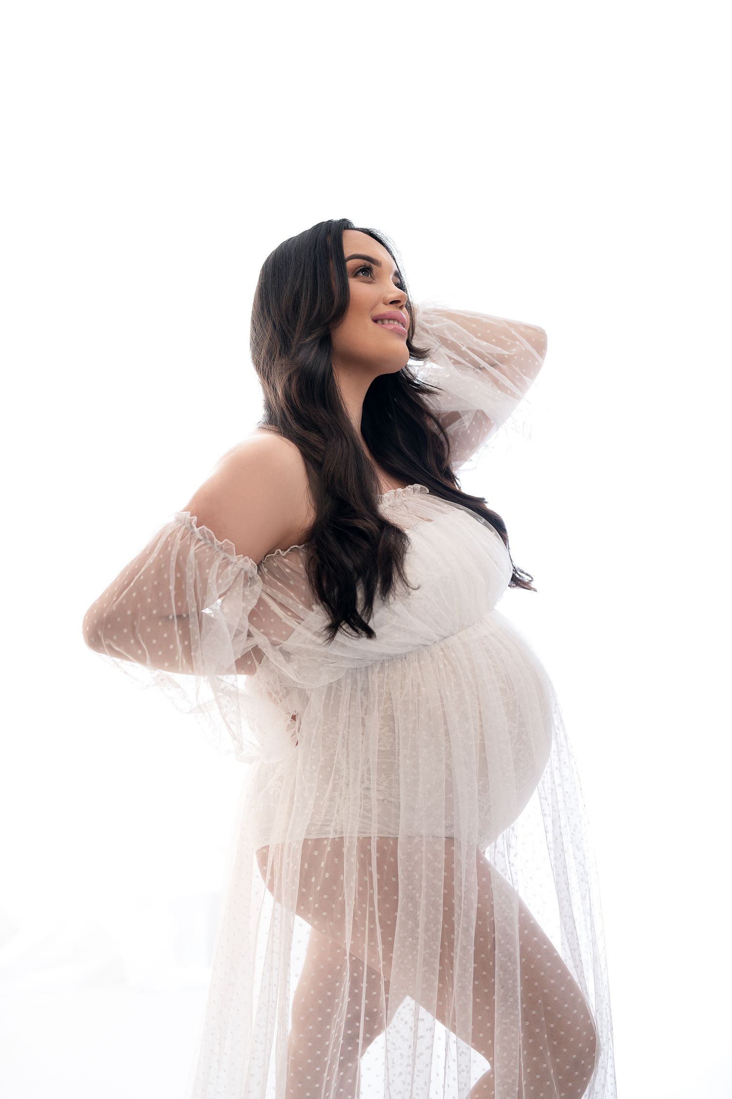 Maternity photo of woman in sheer white dress showing off baby bump against white background.