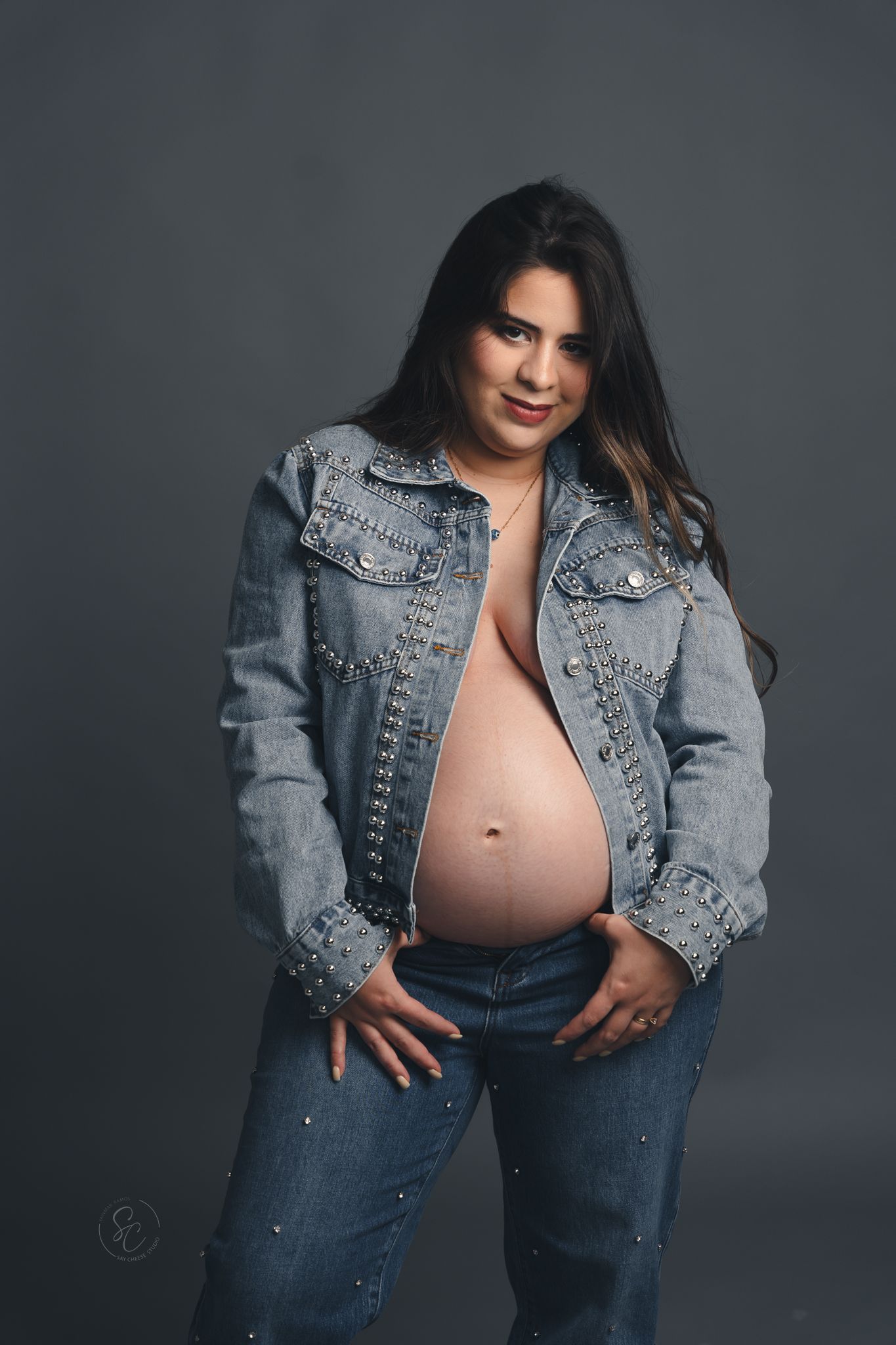 Maternity photoshoot featuring a studded denim jacket and jeans against a dark studio background.