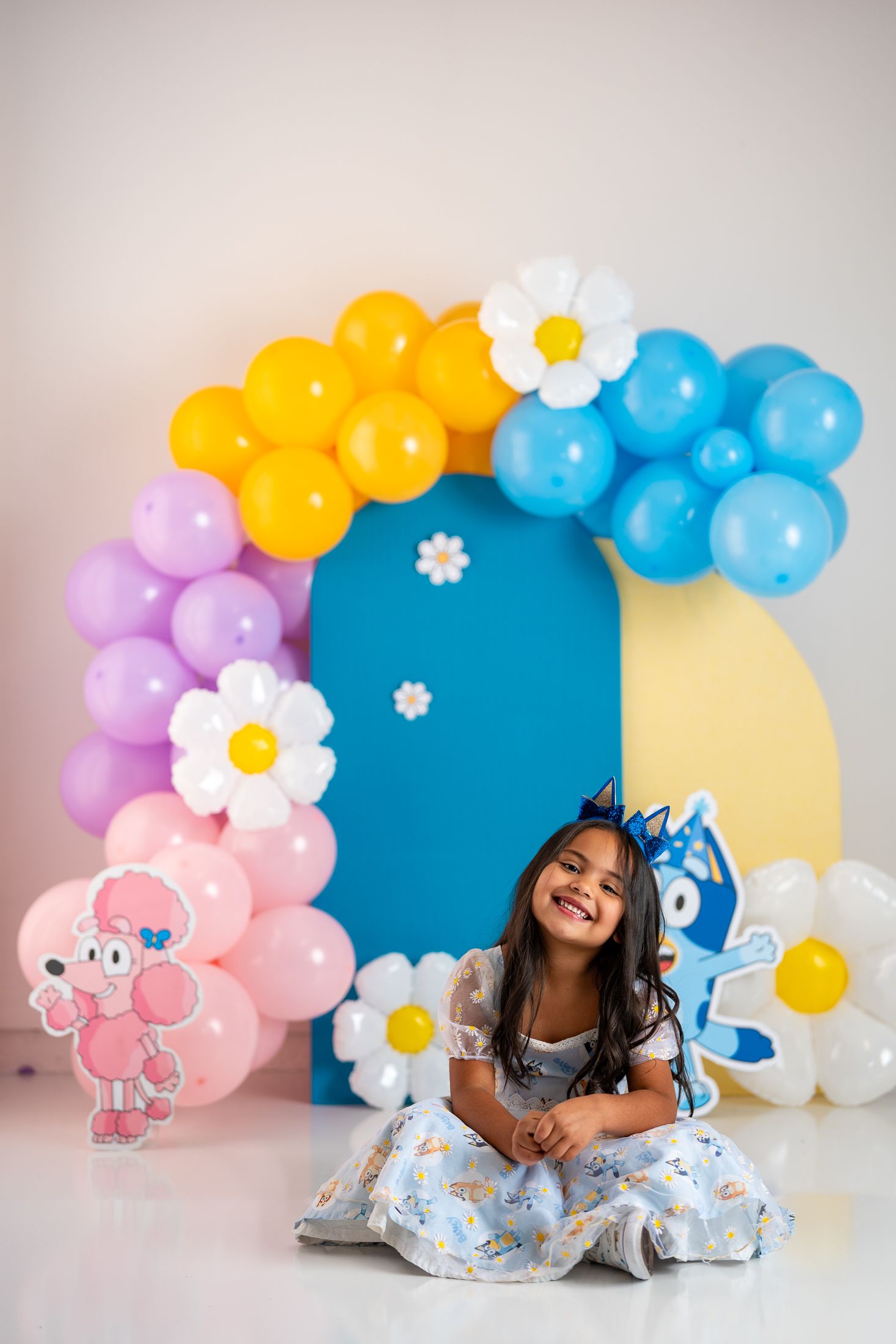 Colorful balloon arch with daisies frames playful scene against pastel backdrop.