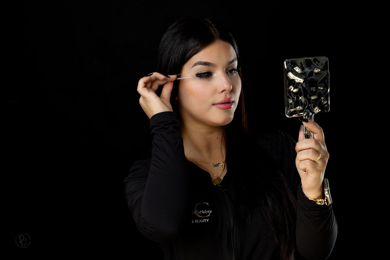 Woman in black outfit poses with beauty tools against dark backdrop.
