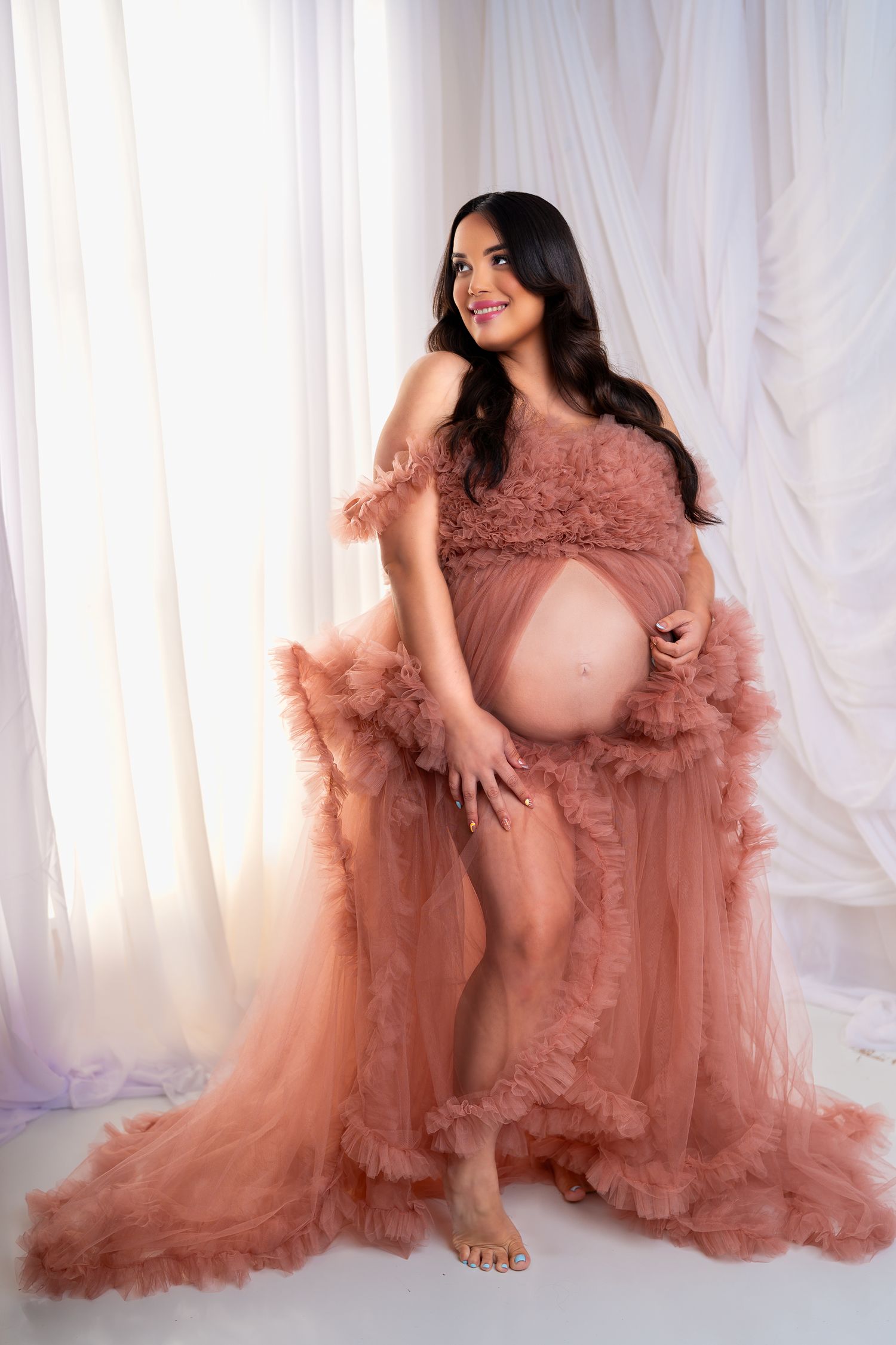 Maternity portrait session with flowing dusty rose tulle gown against white backdrop.