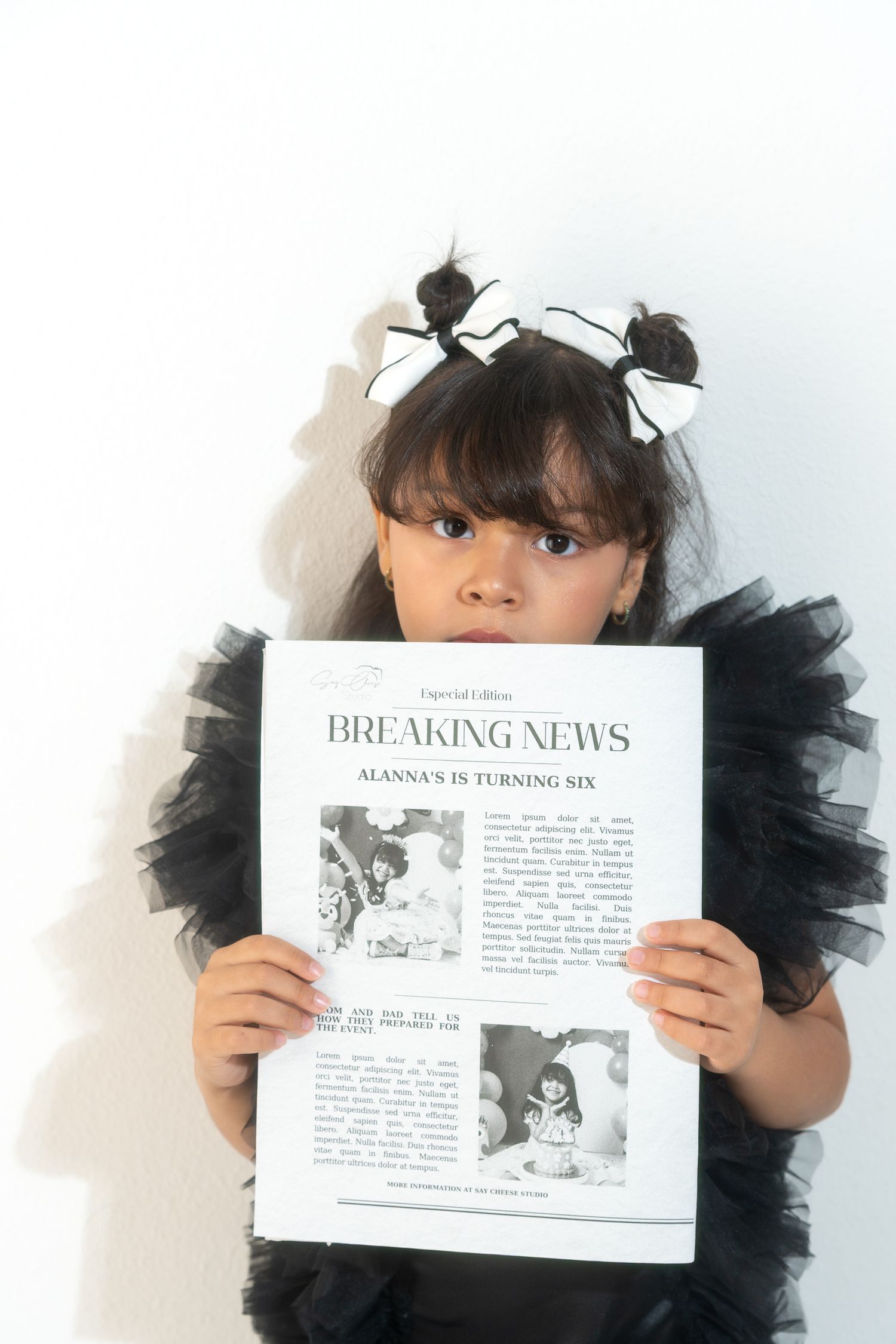 Child holding newspaper-style announcement prints in black tulle dress against white background.