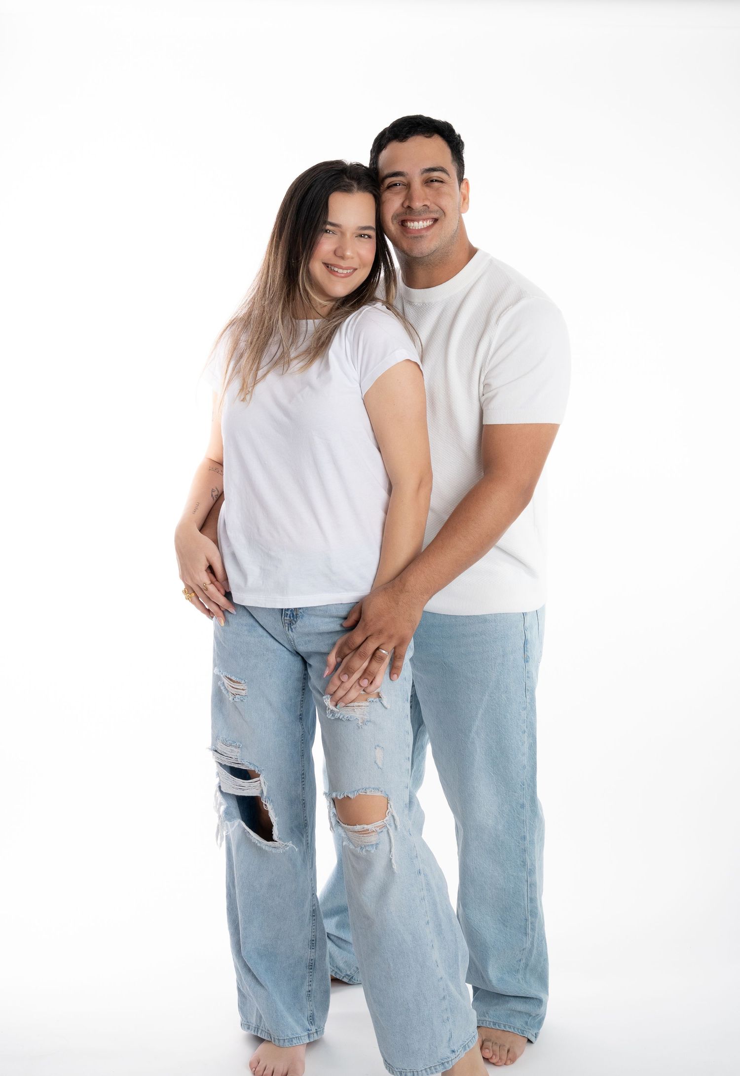 A couple in coordinated white t-shirts and distressed jeans pose intimately against a white backdrop.