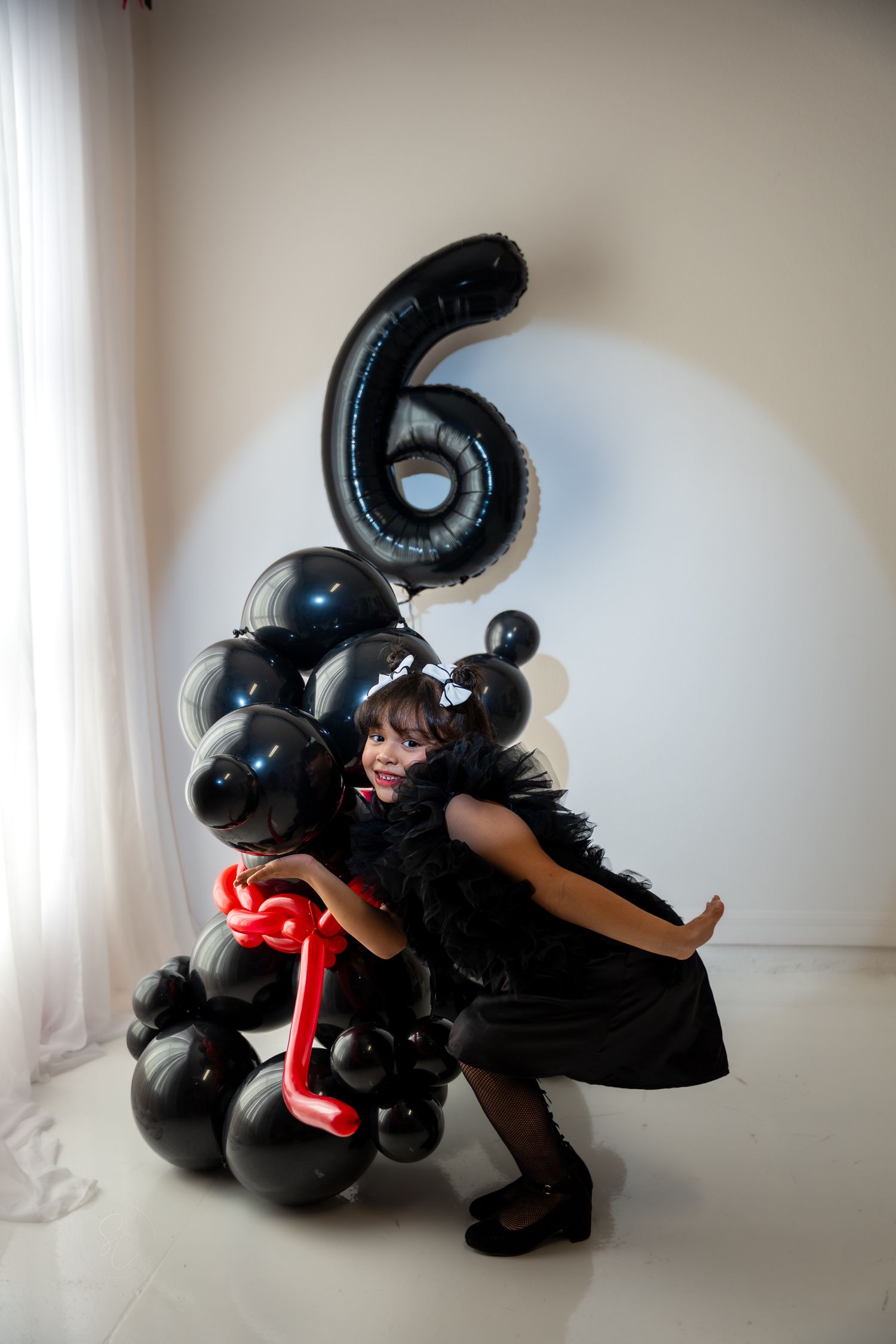Girl in a black feathered dress poses beside a black balloon sculpture with a giant number 6 balloon for a sixth birthday photoshoot.