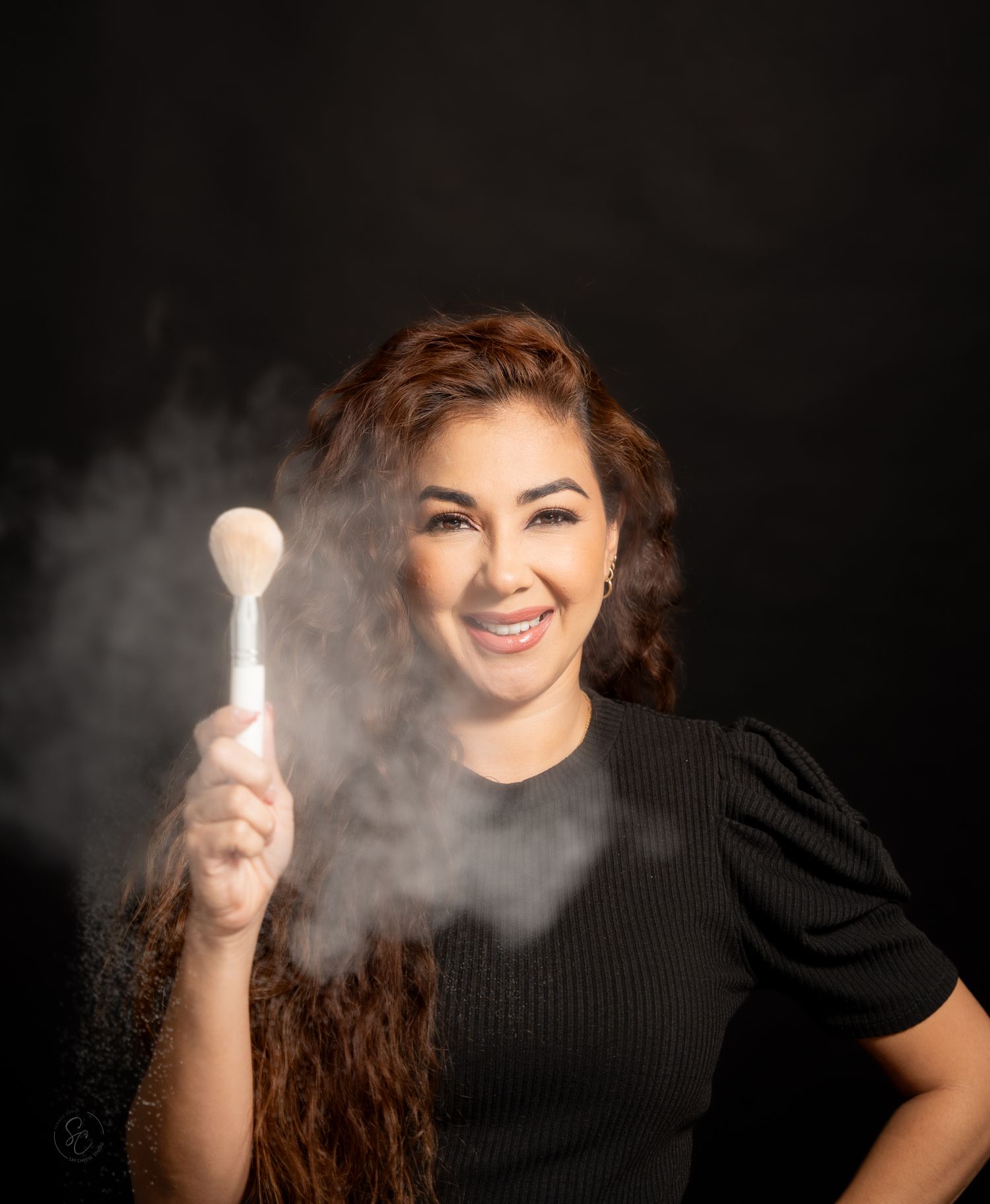 Person in black top with long wavy hair poses playfully against dark background with smoke effects.