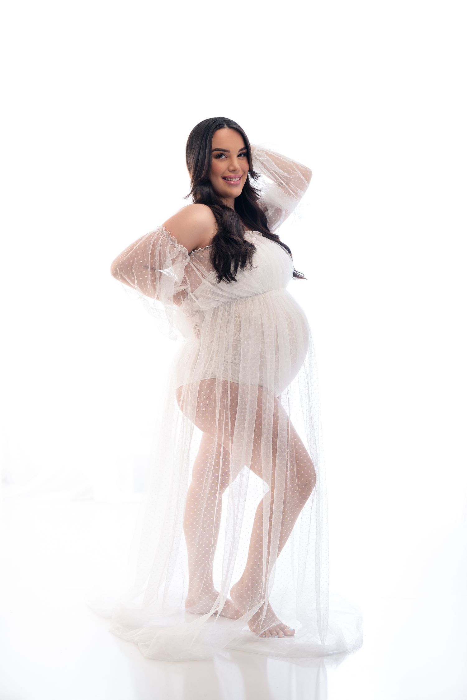 Maternity boudoir photo of woman in sheer white dress against white background.