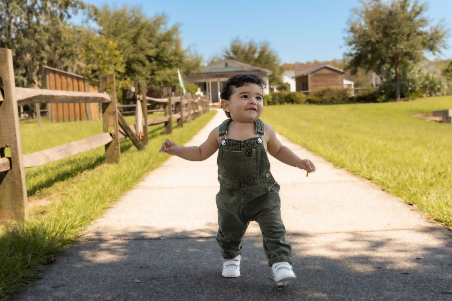 Child walking along sunny path with wooden fence and green grass.