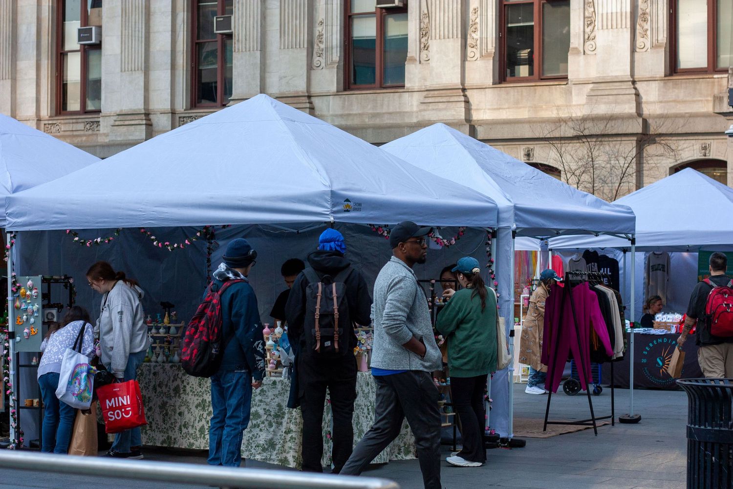 Outdoor market with white canopy tents and shoppers browsing vendor stalls on a city sidewalk.