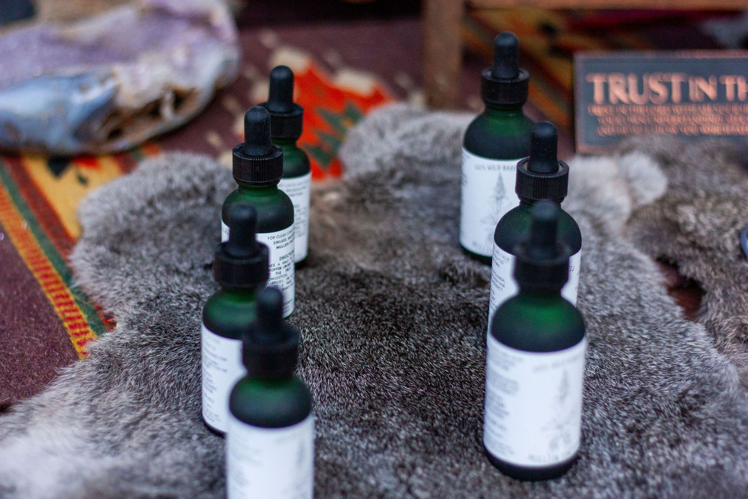 Green dropper bottles with white labels displayed on gray fur at an outdoor market with a Trust sign in background.